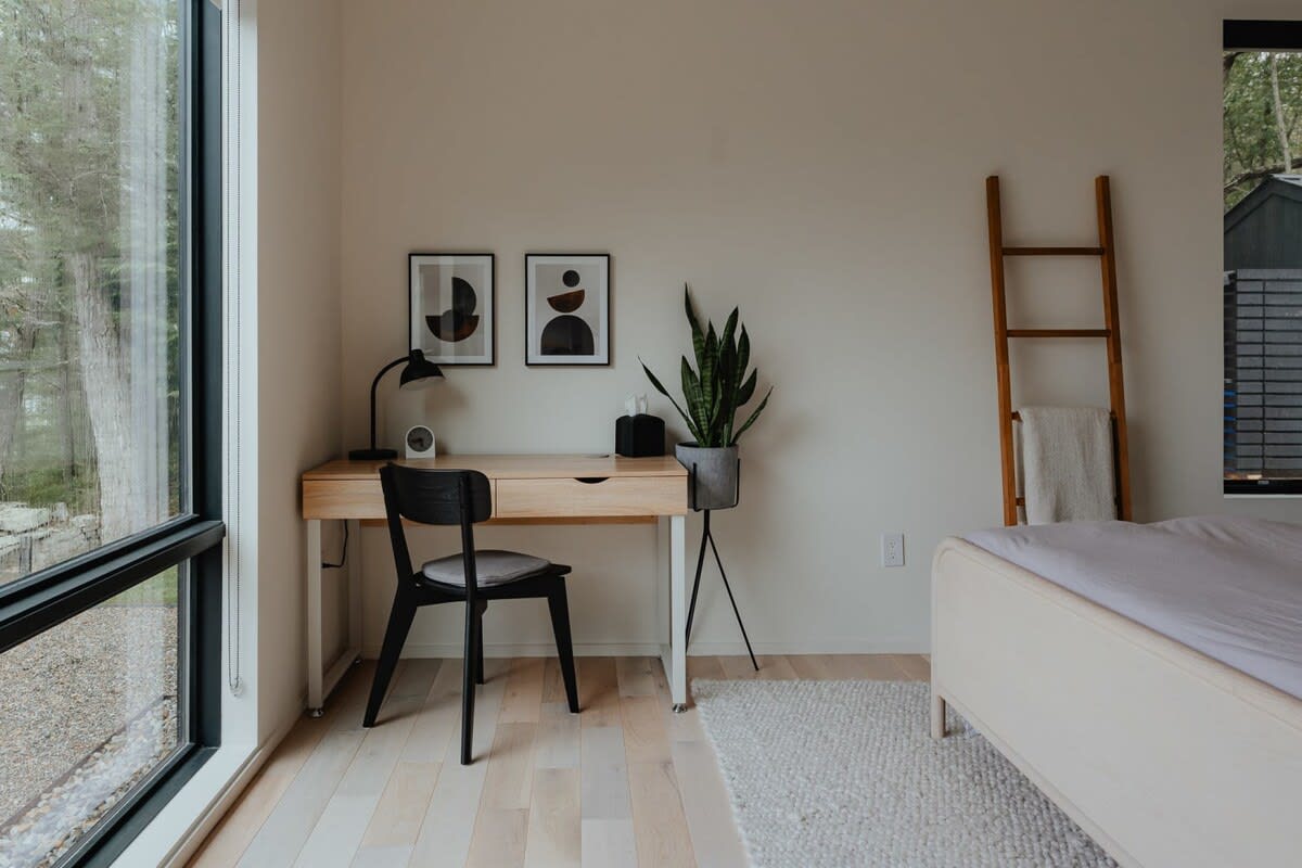 A bright corner of a bedroom features a light wood desk with a black chair, positioned by a large window overlooking the outdoors. Two abstract art prints adorn the wall above the desk, and a tall snake plant in a planter adds a touch of nature.