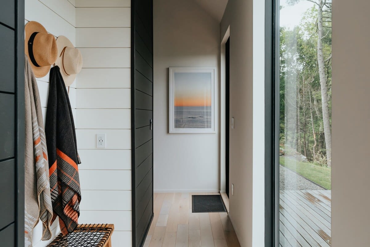 Bright entryway with white shiplap walls, a dark door, and a large window looking out to trees. Hats and towels hang on hooks, and a framed artwork adorns the opposite wall.