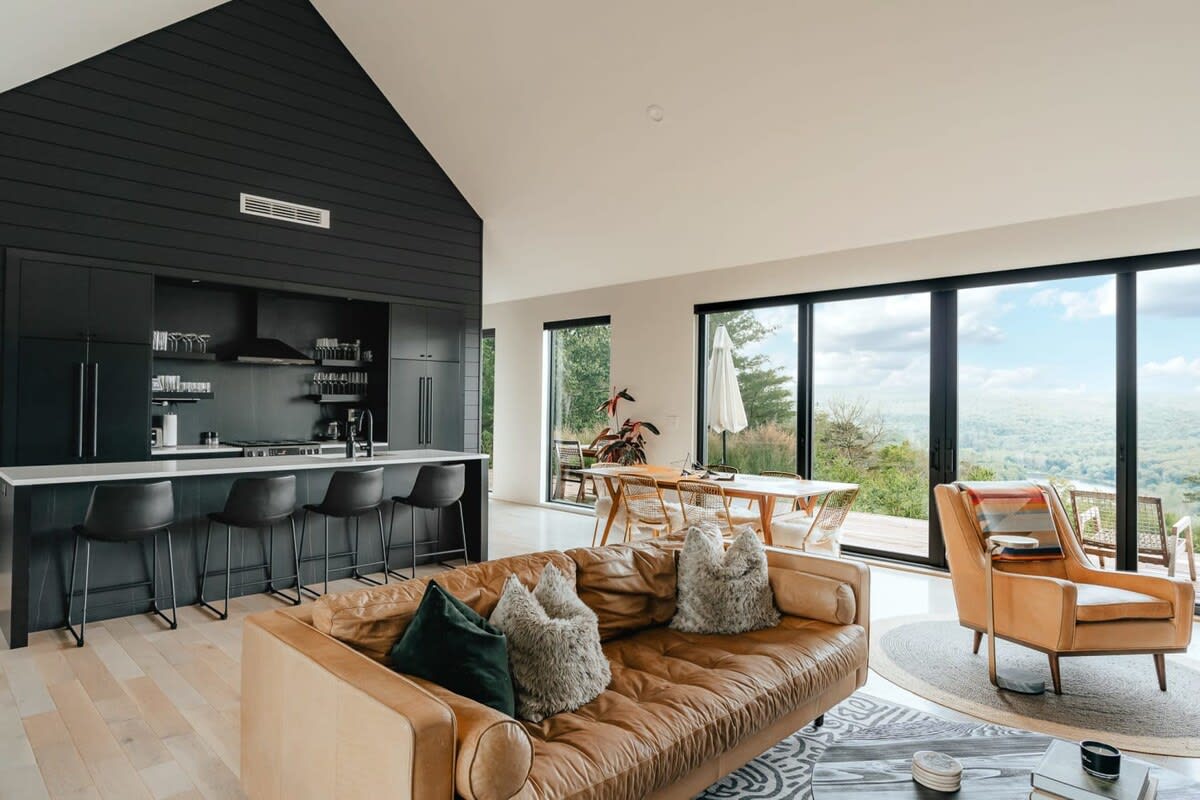 Spacious open-concept living area with a vaulted ceiling, featuring a black kitchen island with bar stools, a brown leather sofa, and a dining area, all illuminated by large windows offering extensive forest views.