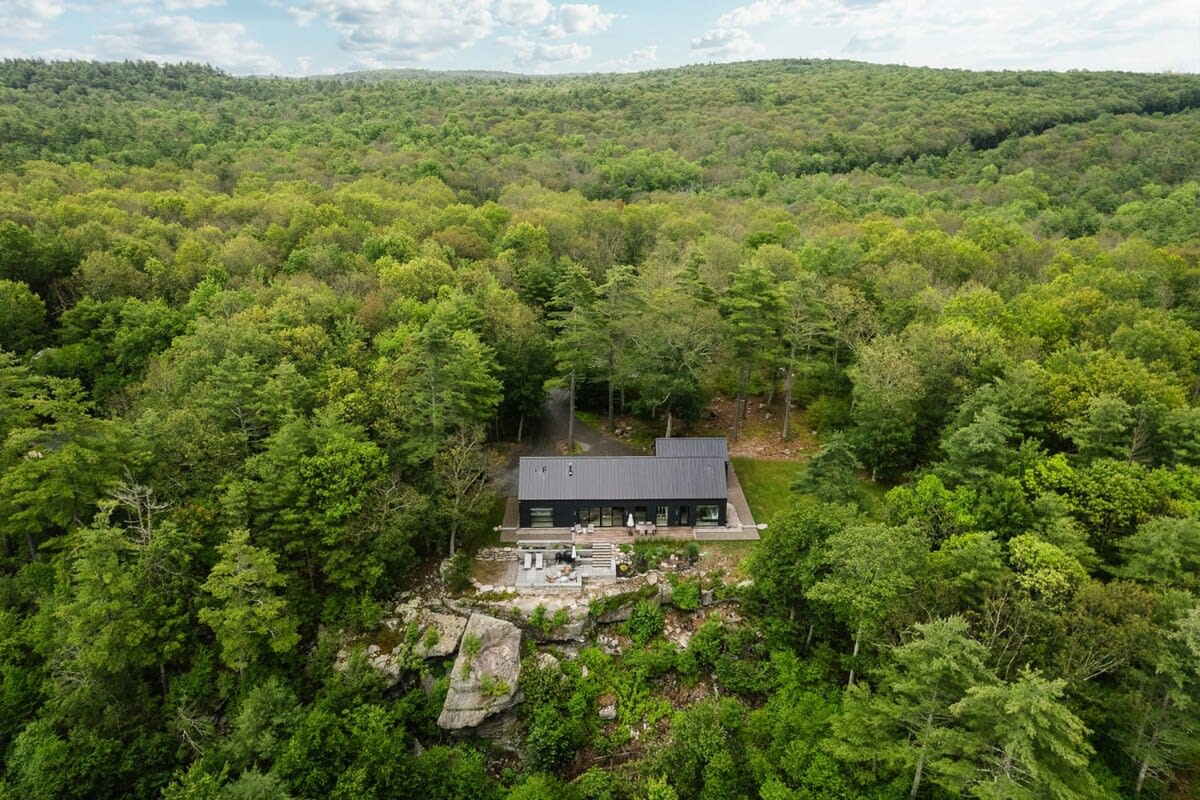 Aerial view of a modern black house with a dark roof, nestled amidst a dense, lush green forest. The house is situated on a cliff edge, offering a secluded and natural setting.