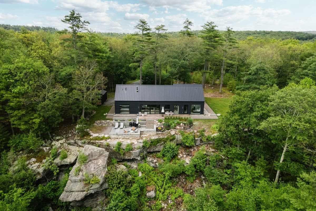Elevated aerial view of a sleek black modern house with a dark metal roof, nestled amidst dense green forest, featuring a multi-level outdoor deck built into a rocky cliffside, offering expansive natural surroundings.