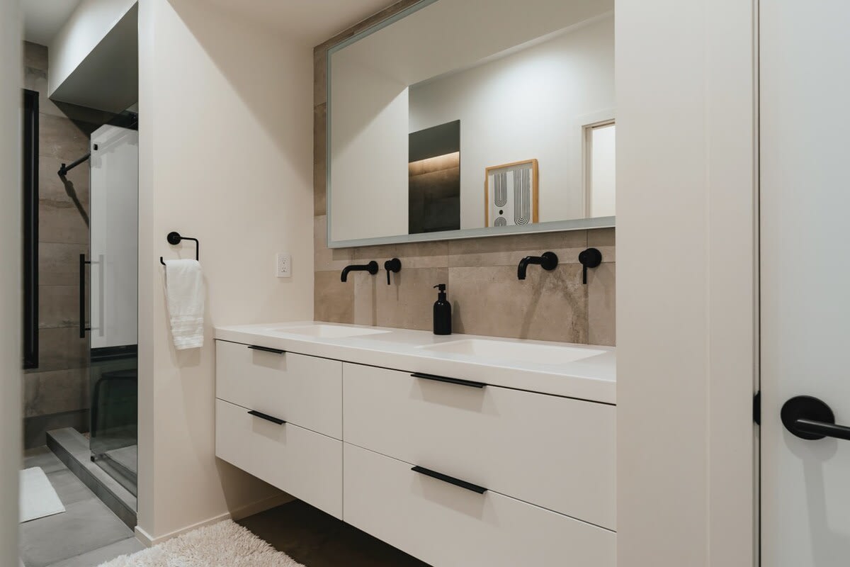 Modern bathroom featuring a double white vanity with black wall-mounted faucets, a large mirror, and a walk-in shower with dark tiles and a glass enclosure.