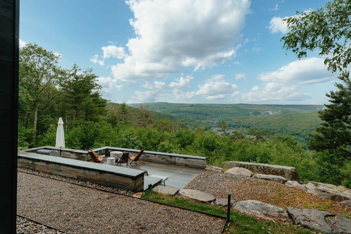 Expansive view from a stone patio overlooking a vast, tree-covered valley and distant hills under a partly cloudy sky, with a small gravel area in the foreground.