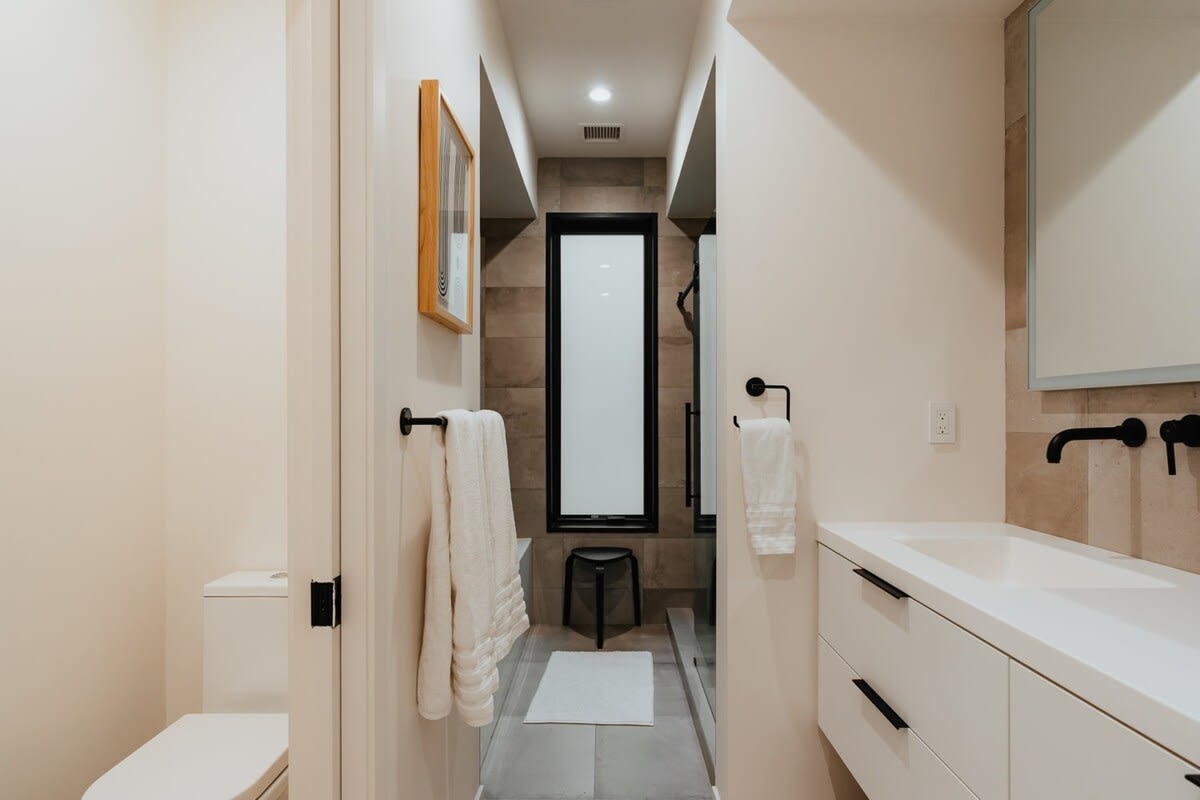 Contemporary bathroom with a white toilet, a black towel bar holding white towels, and a walk-in shower with dark tiled walls and a tall narrow window.