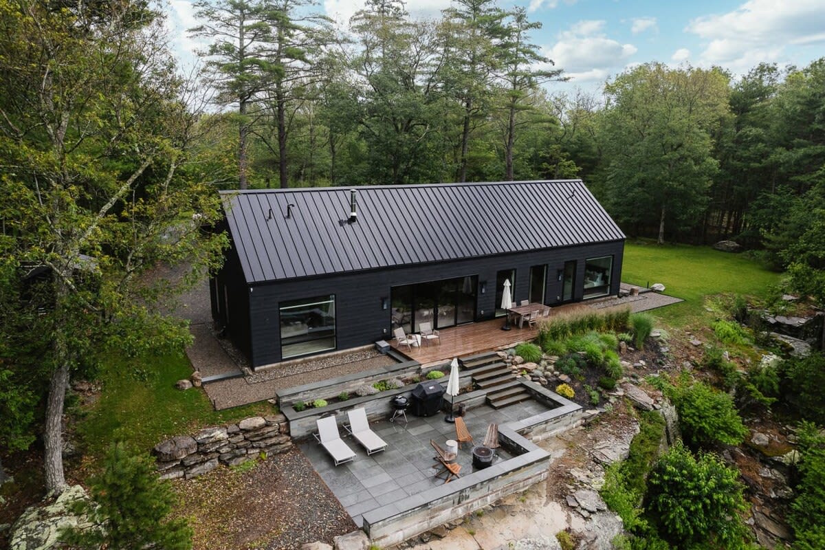 Aerial view of a modern black house with a dark metal roof, surrounded by lush green trees, featuring a multi-level patio with lounge chairs and a fire pit overlooking a valley.