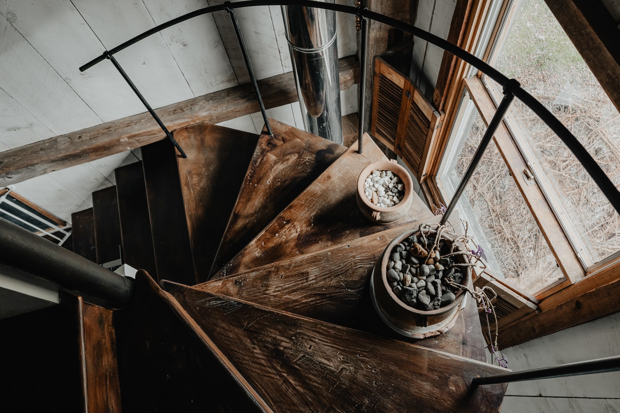 A top-down view of a rustic wooden spiral staircase with a black metal railing, adorned with potted plants filled with pebbles. Natural light streams in from a nearby window.