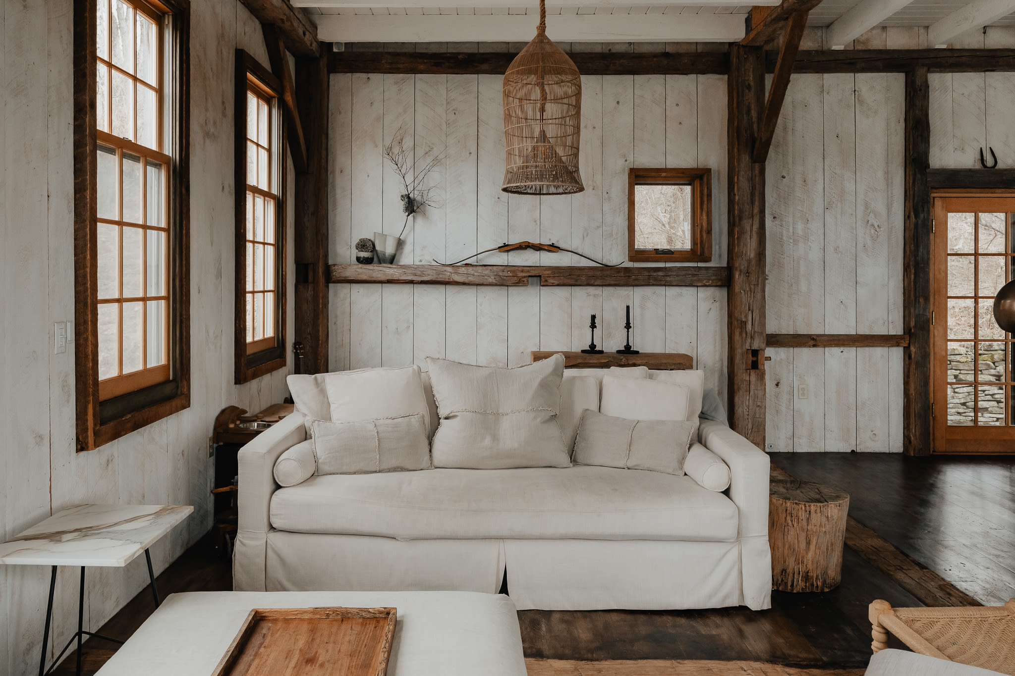 A cozy living room features a white slipcovered sofa with linen pillows, set against white shiplap walls and dark wood beams. A unique woven birdcage-style pendant light hangs from the ceiling, and a rustic wooden shelf displays a bow and arrows.