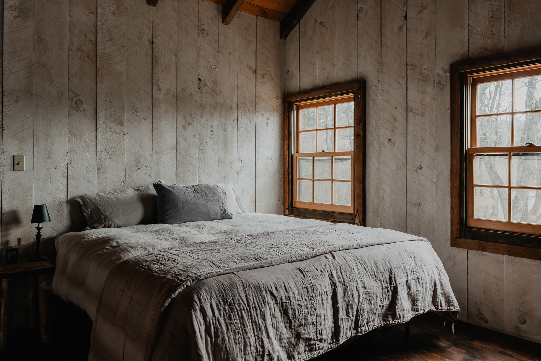 A cozy loft bedroom with a bed dressed in grey bedding, flanked by two windows with wooden frames. The room features white shiplap walls and a dark wood floor.