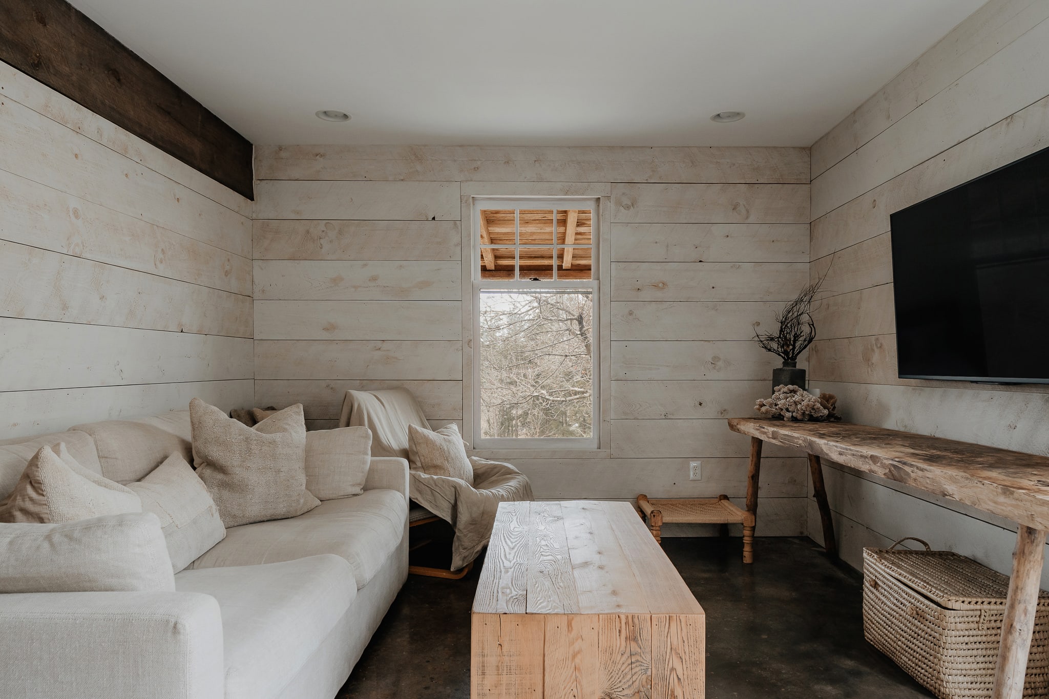 A bright living room with white shiplap walls and dark concrete floors features a large cream-colored sectional sofa and a rustic wooden coffee table. A window offers a glimpse of the outdoors, and a wall-mounted TV is visible on the right.