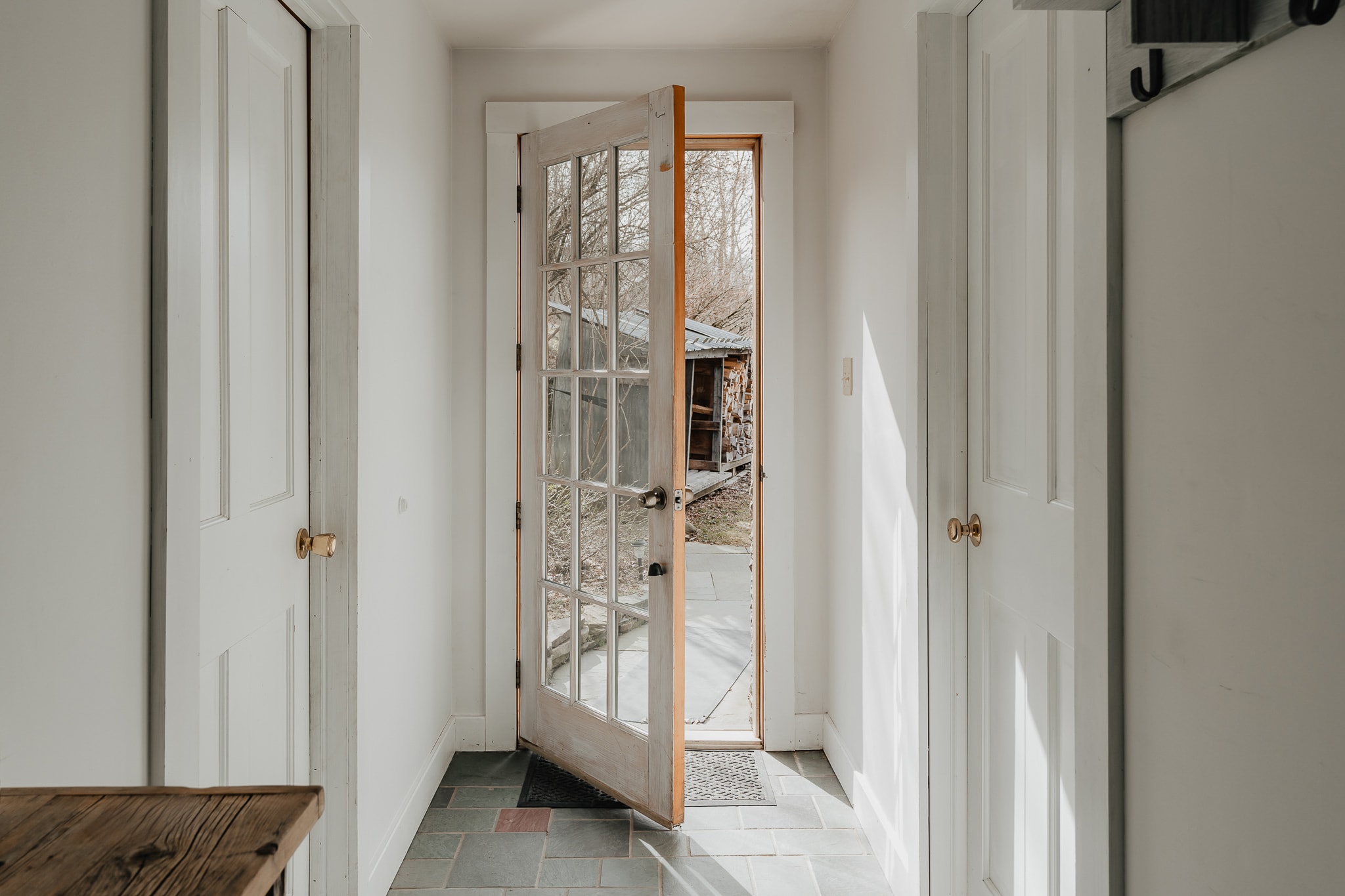 A bright entryway with white walls and slate tile flooring features an open glass-paned door leading to the exterior. Two white doors flank the entrance, and sunlight streams in, highlighting the simple, clean design.