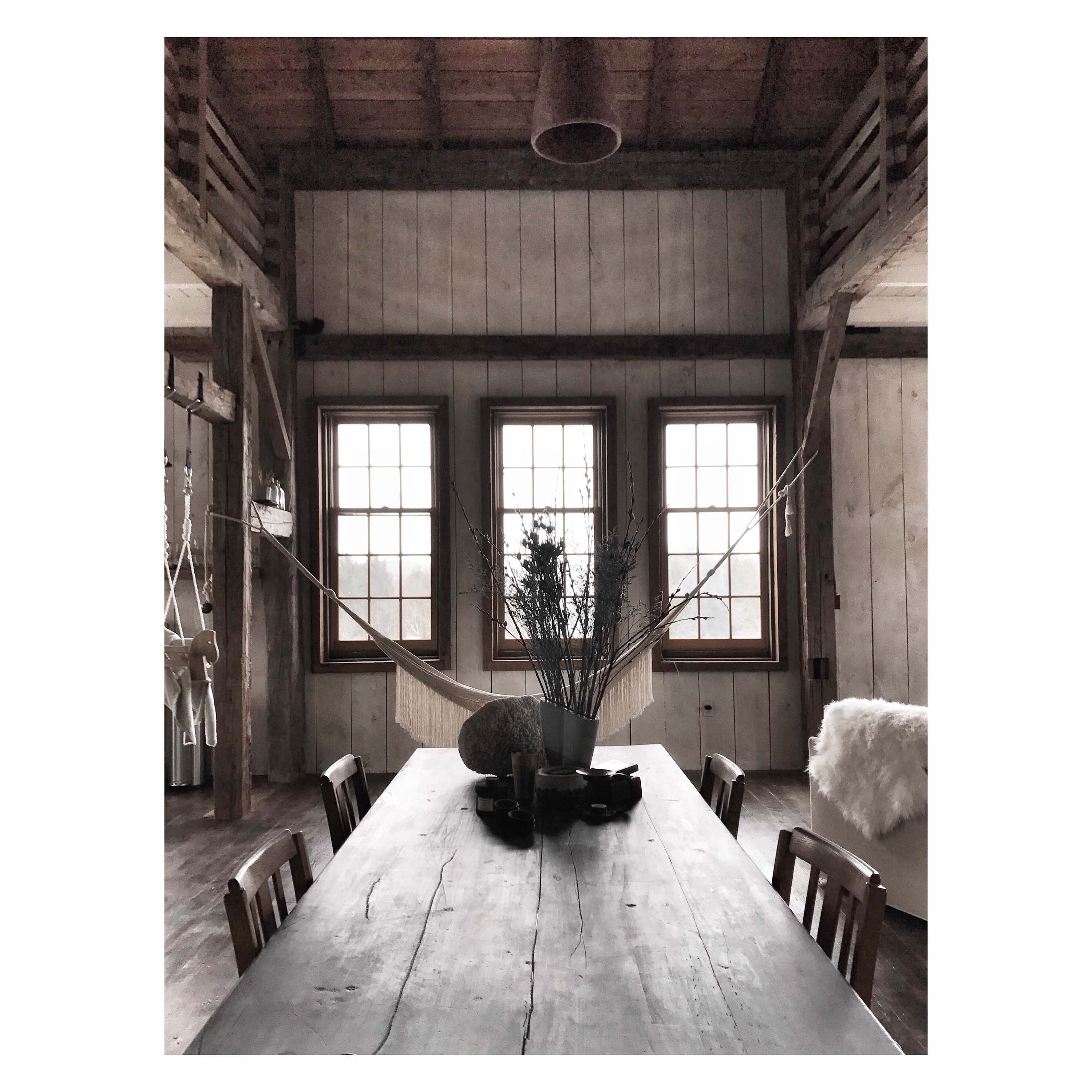 A rustic dining room features a long wooden table with chairs, positioned in front of three large windows. A hammock is visible hanging in the background, and the room has exposed wood beams and shiplap walls.