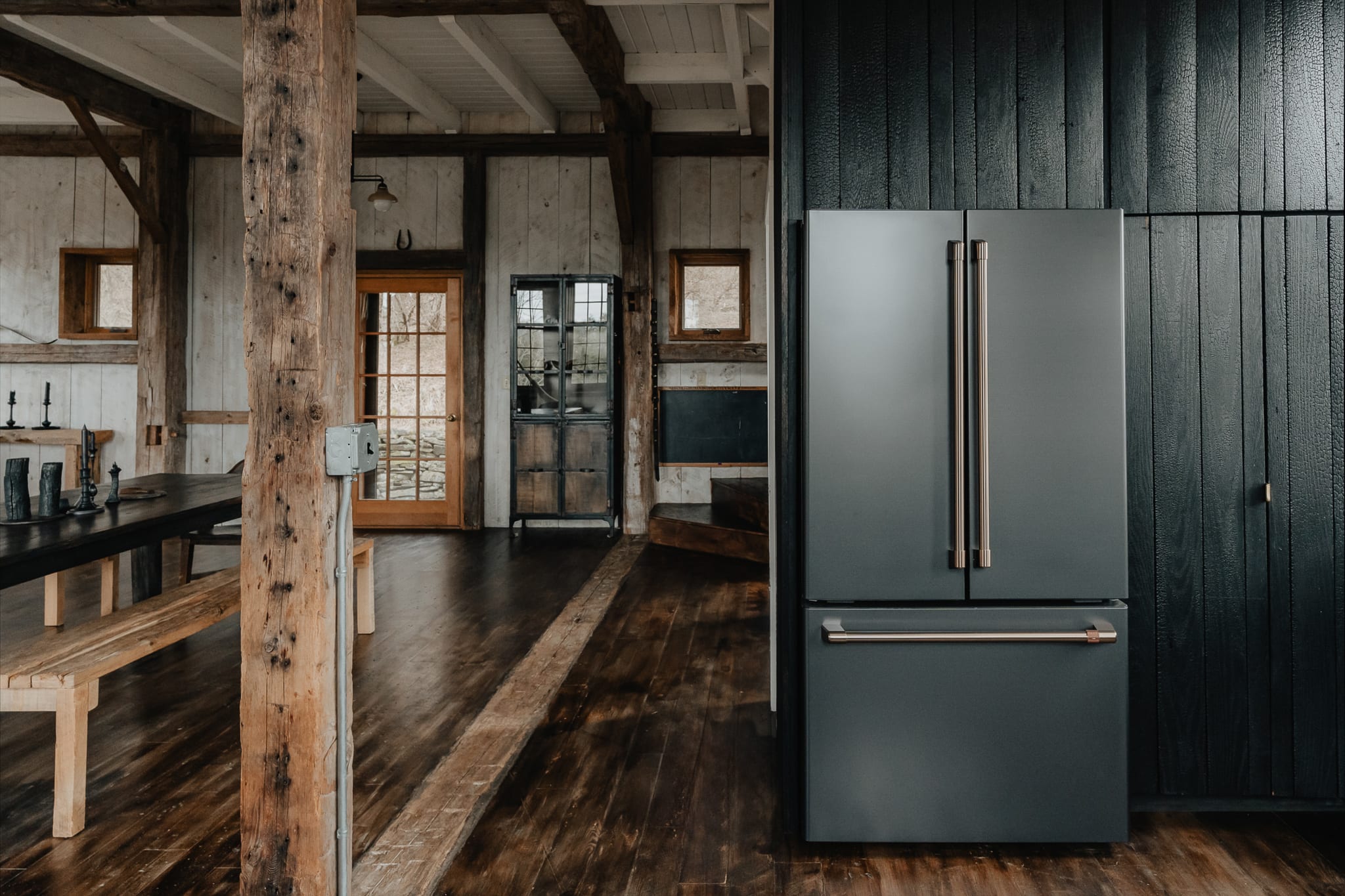 A modern matte black refrigerator stands against dark shiplap paneling, contrasting with the rustic white shiplap walls and exposed wood beams of the open-concept space. Part of the dining area is visible in the background.