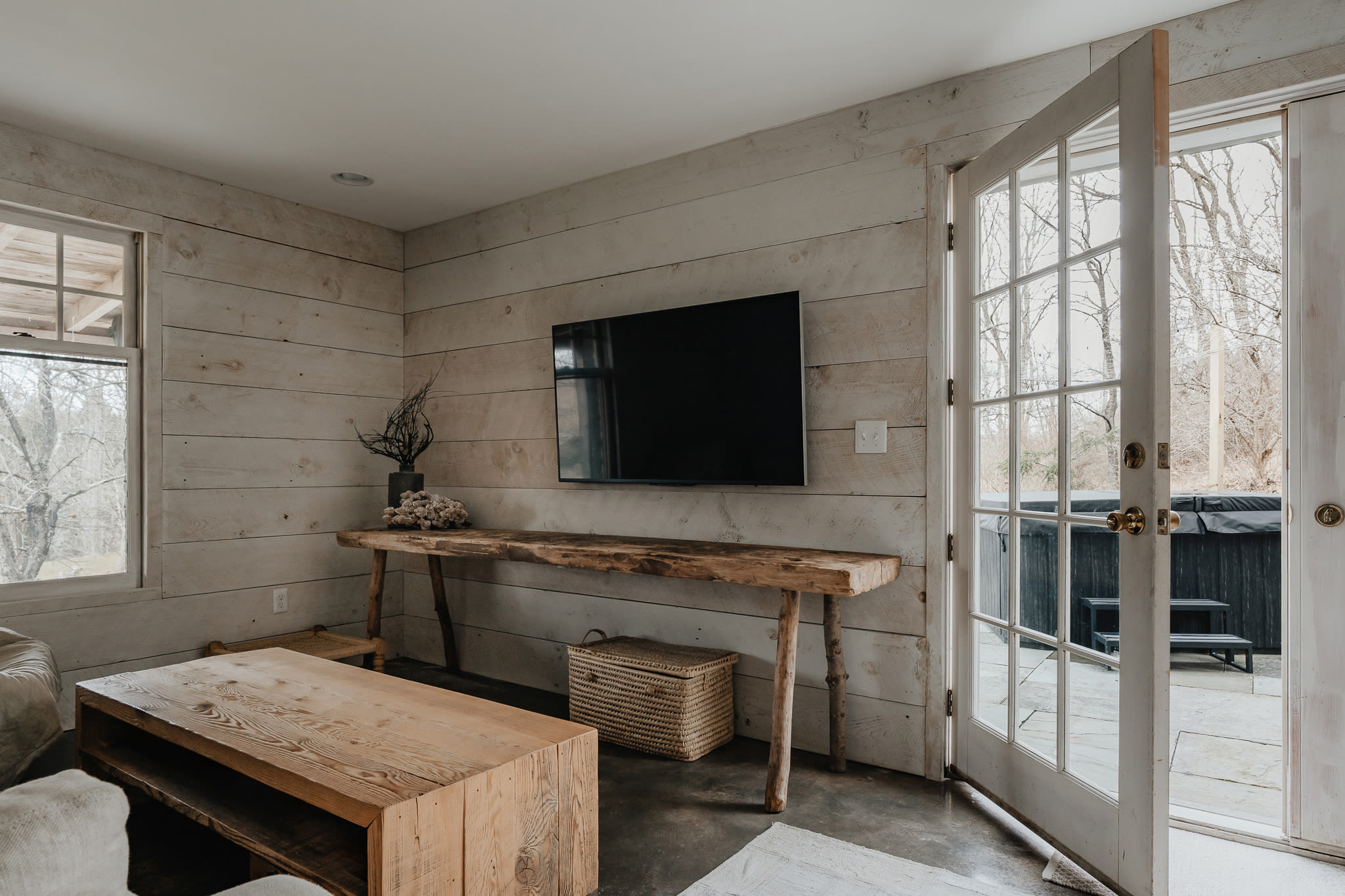A living area with white shiplap walls, a wall-mounted TV above a long, rustic wooden console table, and a French door opening to an outdoor patio with a hot tub.