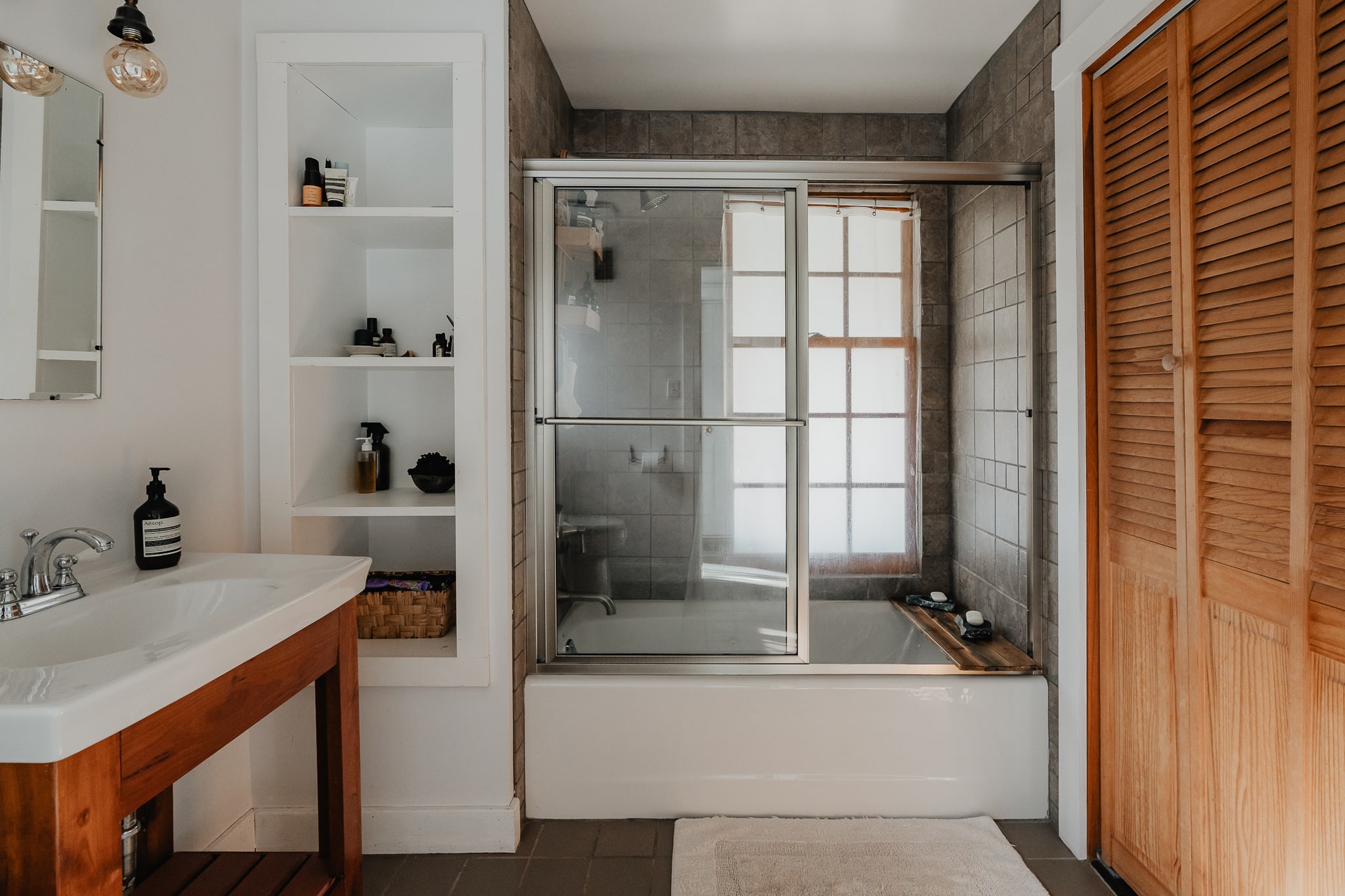 A modern bathroom featuring a white vanity with a rectangular sink, built-in open shelving with toiletries, and a shower-tub combo with sliding glass doors and dark tiled walls.