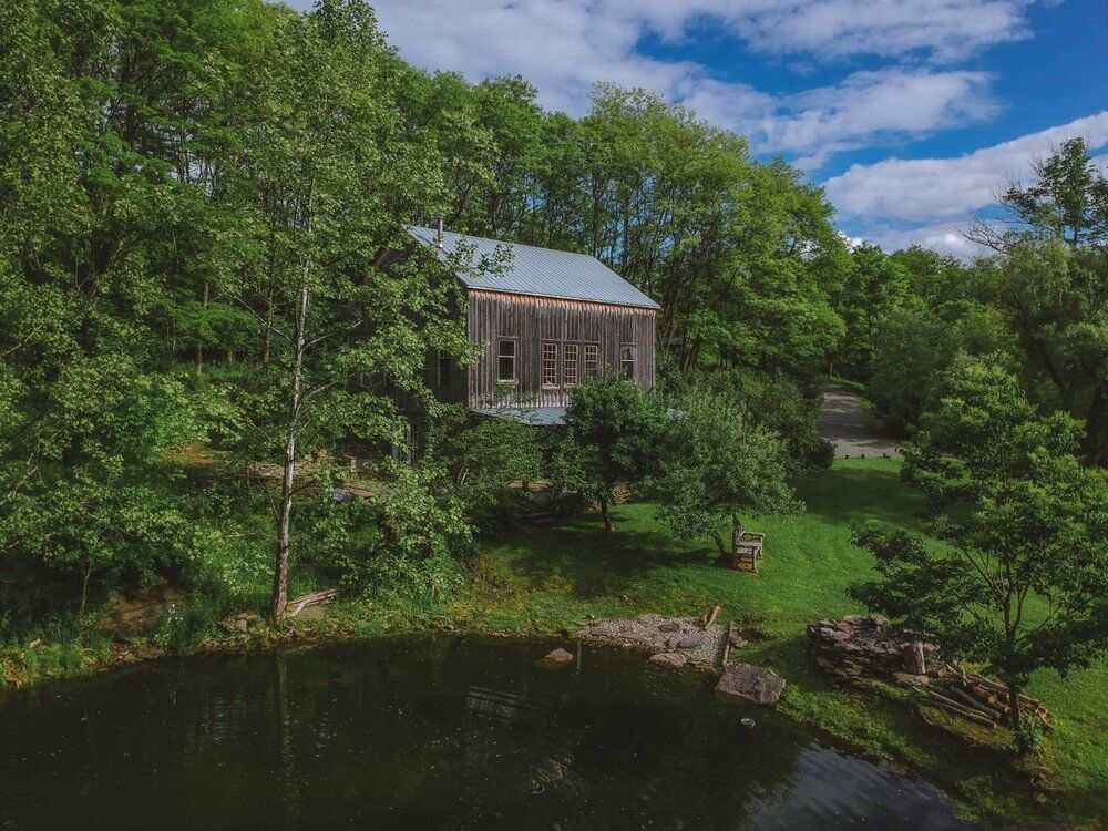 An elevated view of a rustic barn-style house with wooden siding and a metal roof, nestled among lush green trees beside a dark pond. The property is surrounded by natural landscape under a partly cloudy sky.