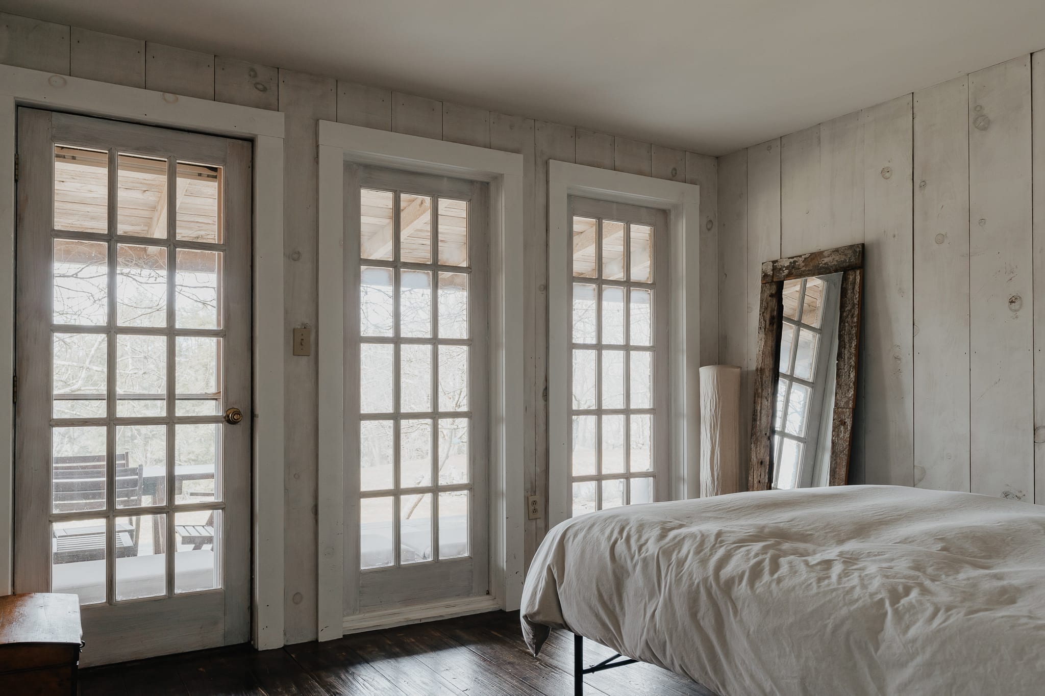 A bright bedroom with white shiplap walls, dark wood floors, and three large French doors letting in natural light. A bed with white bedding and a tall, rustic mirror lean against the wall.