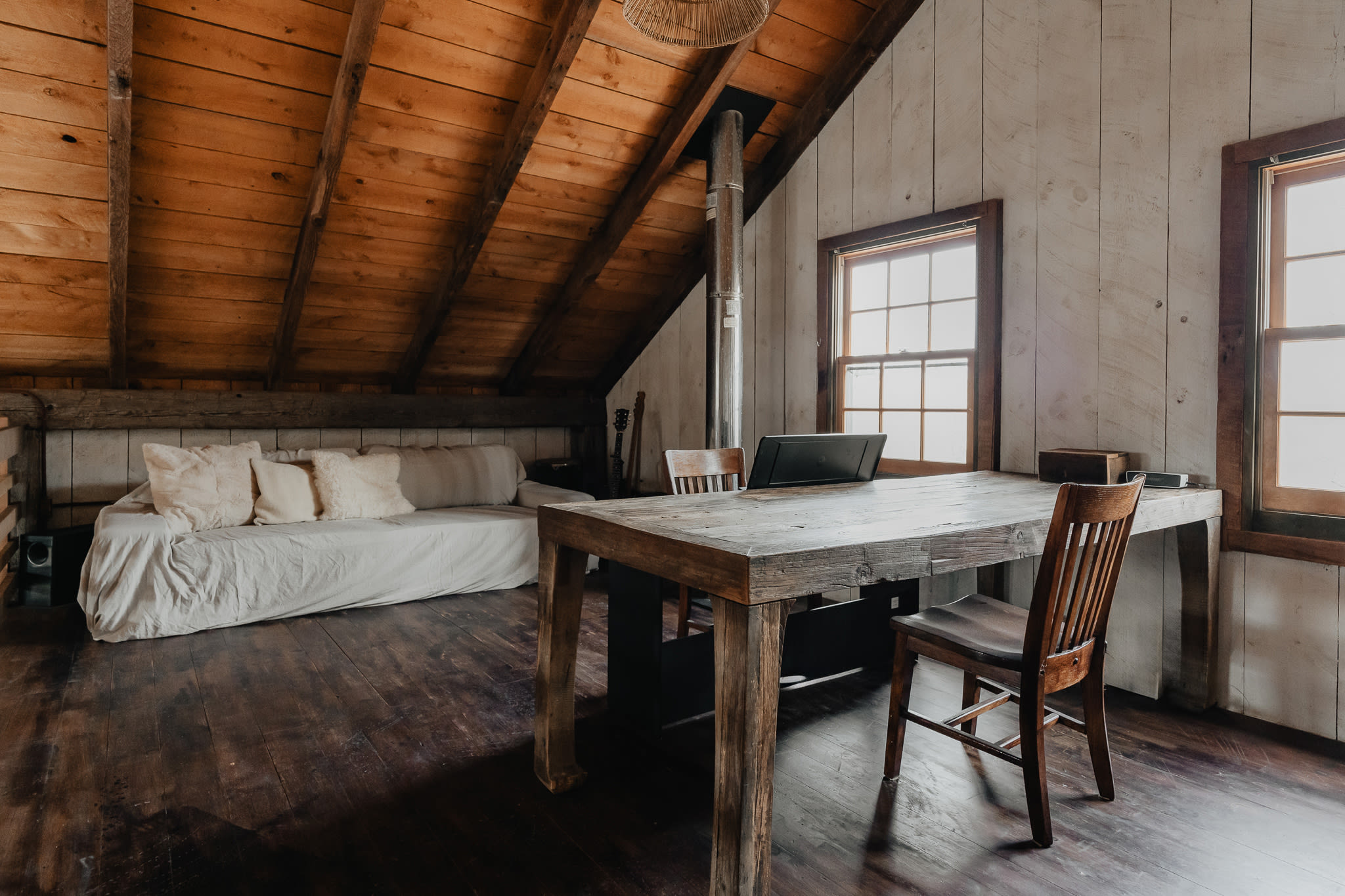 A spacious loft area with a sloped wooden ceiling and white shiplap walls, featuring a daybed with pillows and a large wooden desk with chairs, illuminated by natural light from windows.