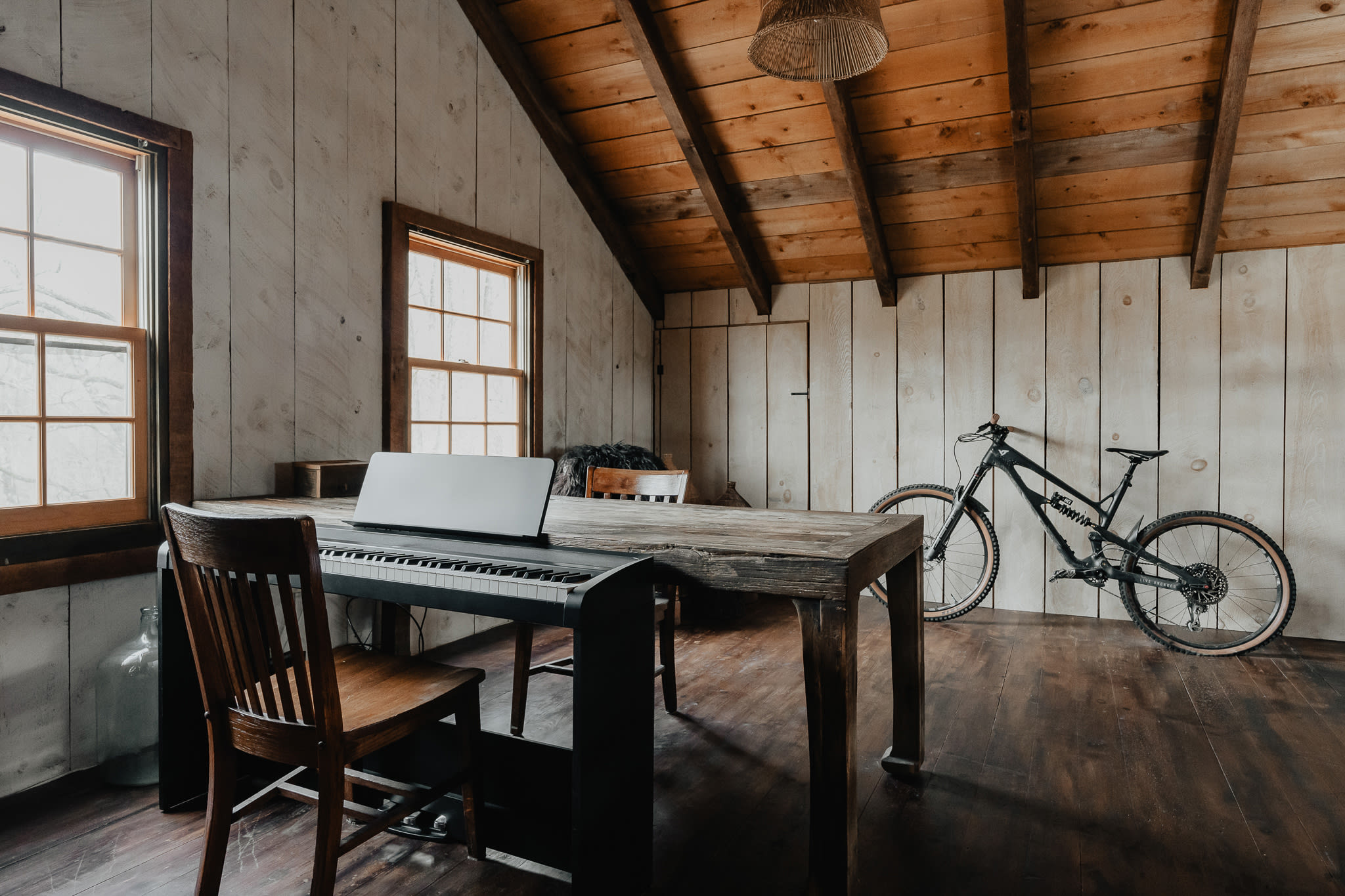 A rustic attic room with sloped wooden plank walls and ceiling, featuring a large wooden desk with a digital piano and a wooden chair. A mountain bike is propped against the far wall, and two windows provide natural light.