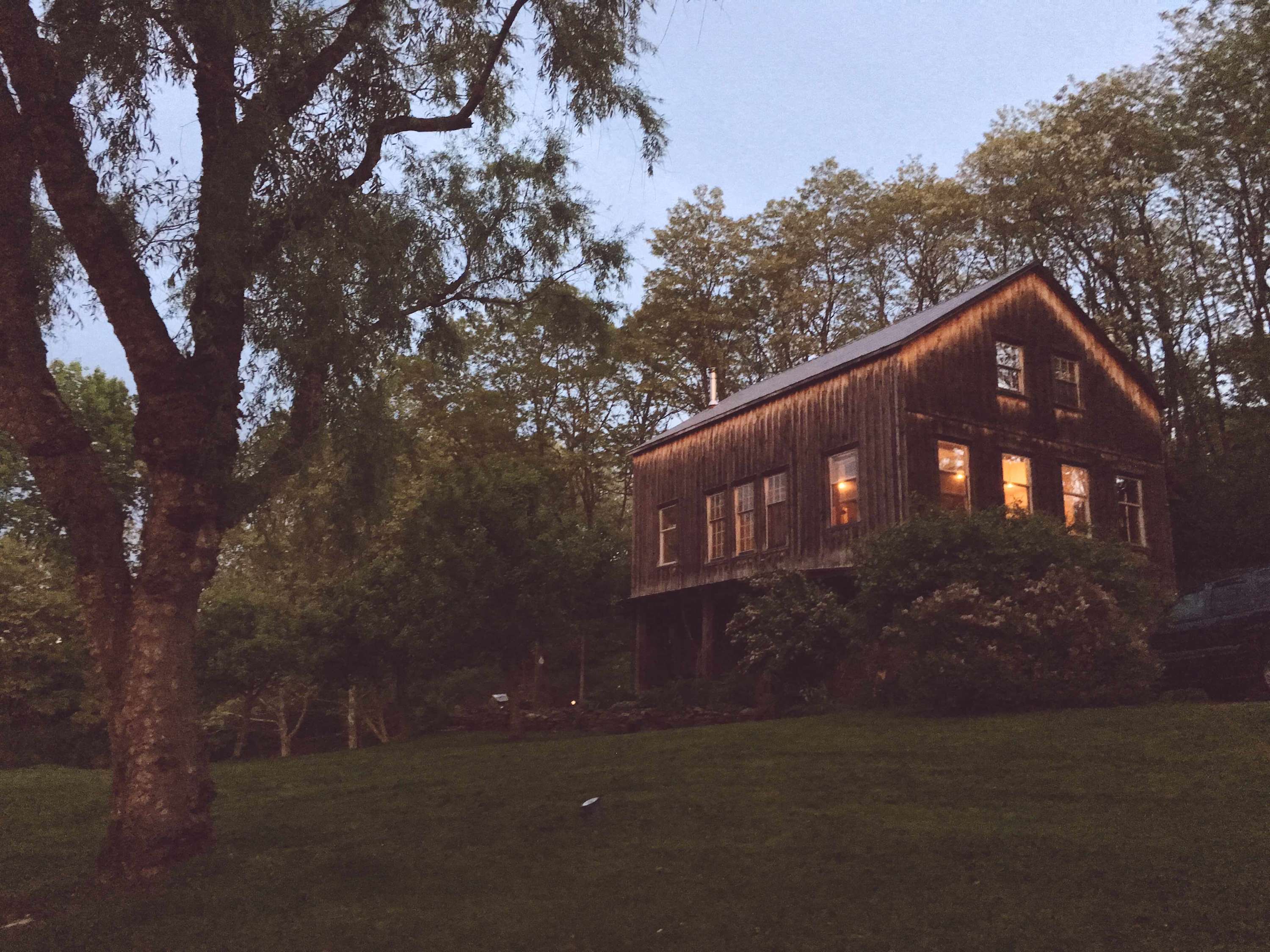 A charming rustic barn-style house with dark wooden siding and multiple windows glowing warmly from within, set against a twilight sky. The house is surrounded by trees and a grassy lawn, creating a cozy evening ambiance.