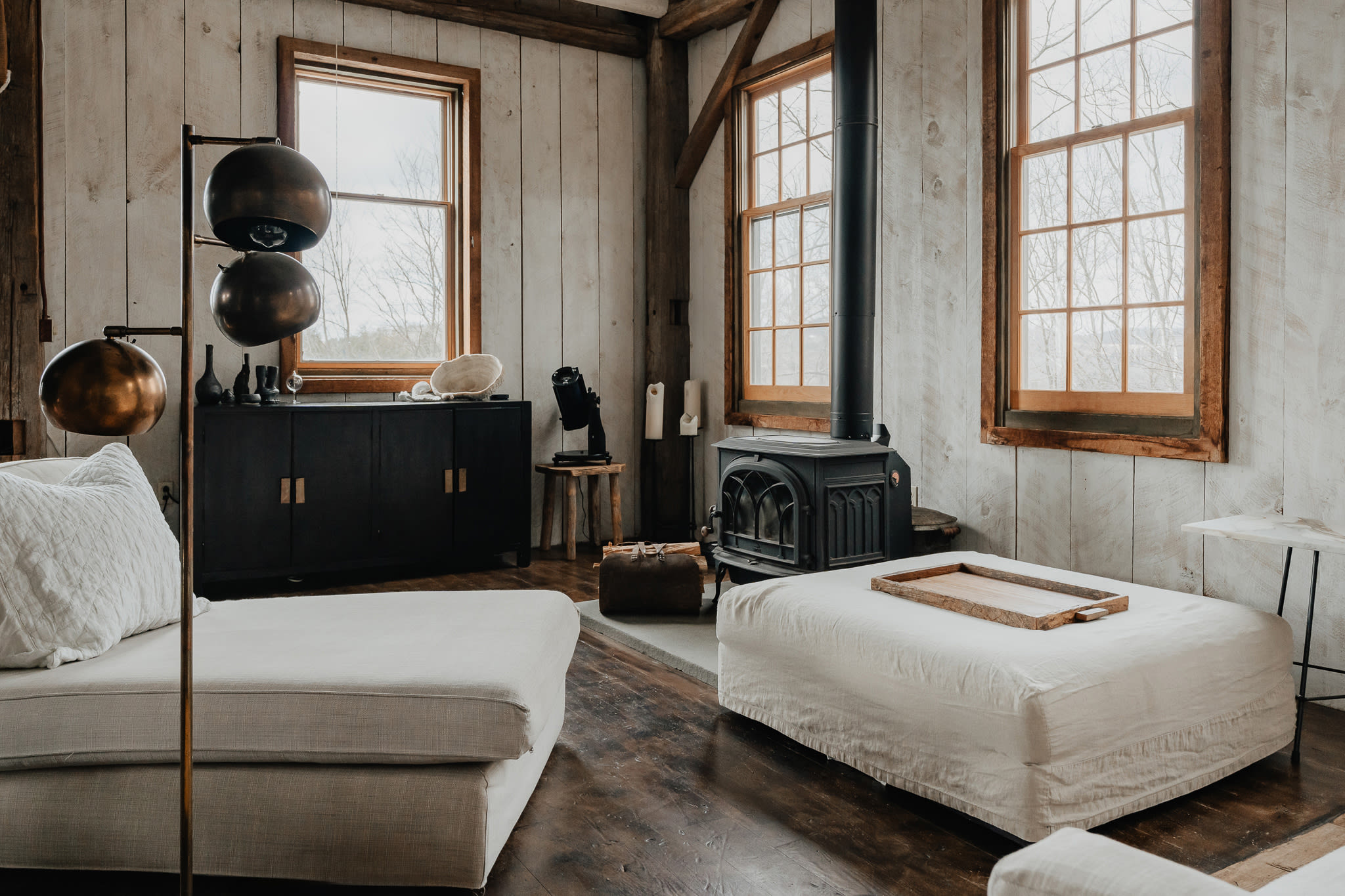 A rustic living space with a black wood-burning stove as the focal point, surrounded by white shiplap walls and dark wood floors. Two light-colored chaises face each other, and large windows offer views of the outdoors.