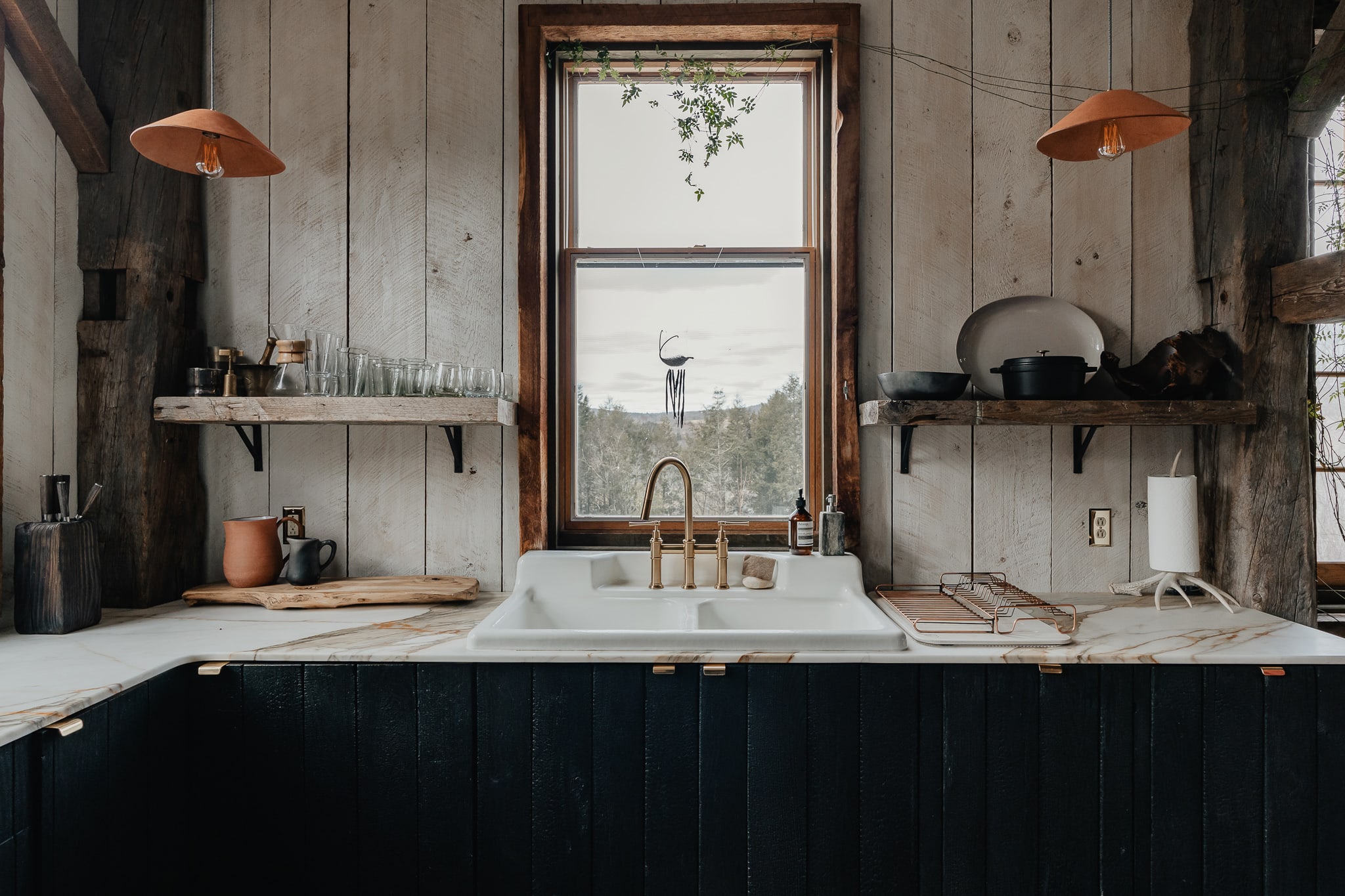 A rustic kitchen features a white farmhouse sink with a brass faucet, set into a marble-patterned countertop. Dark shiplap cabinets are below, and open wooden shelves display dishes, all against white shiplap walls.