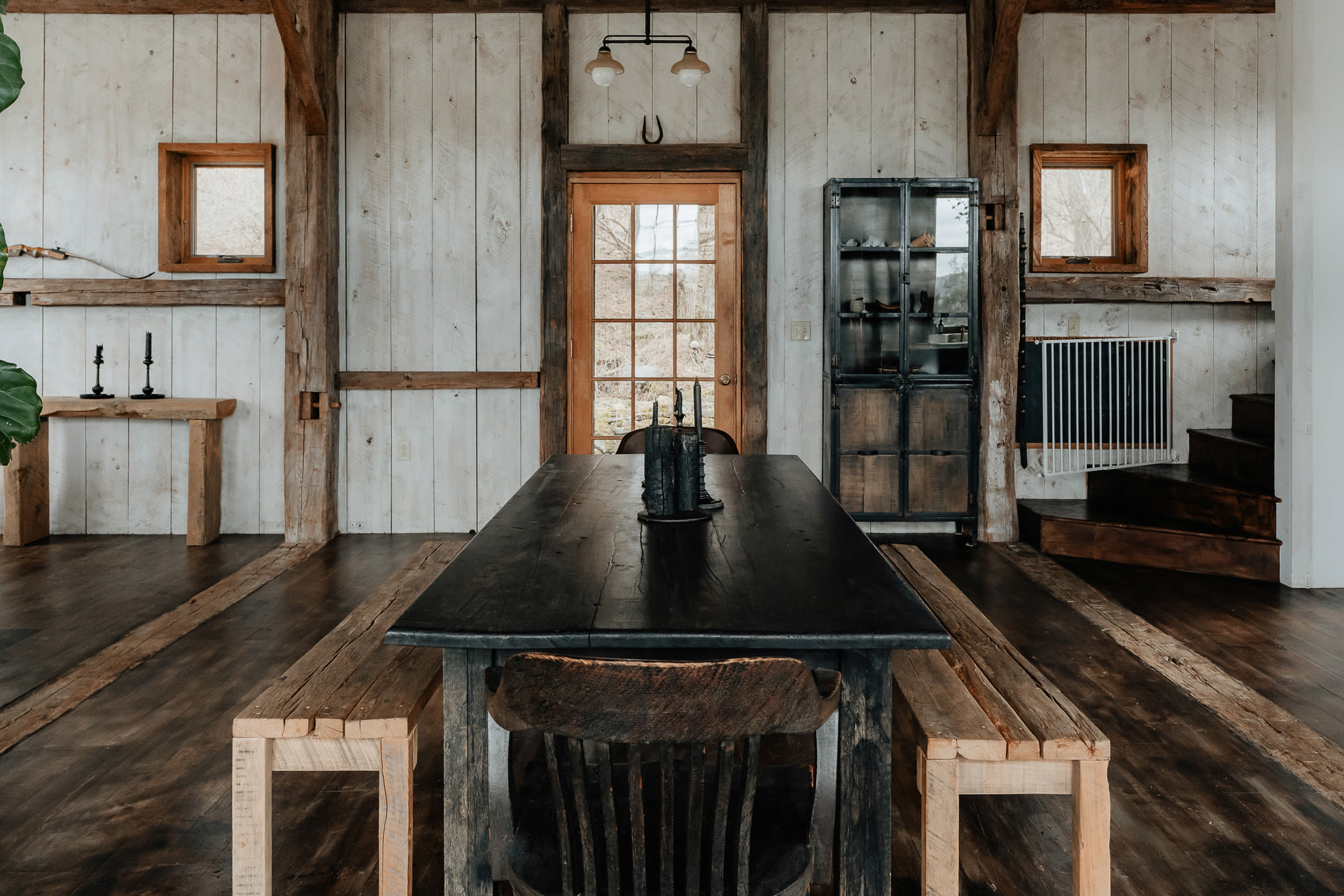 A spacious dining area features a long, dark wooden table with matching benches and a single chair, set against white shiplap walls and exposed wood beams. A glass-paned door leads outside, and a dark metal cabinet stands in the corner.
