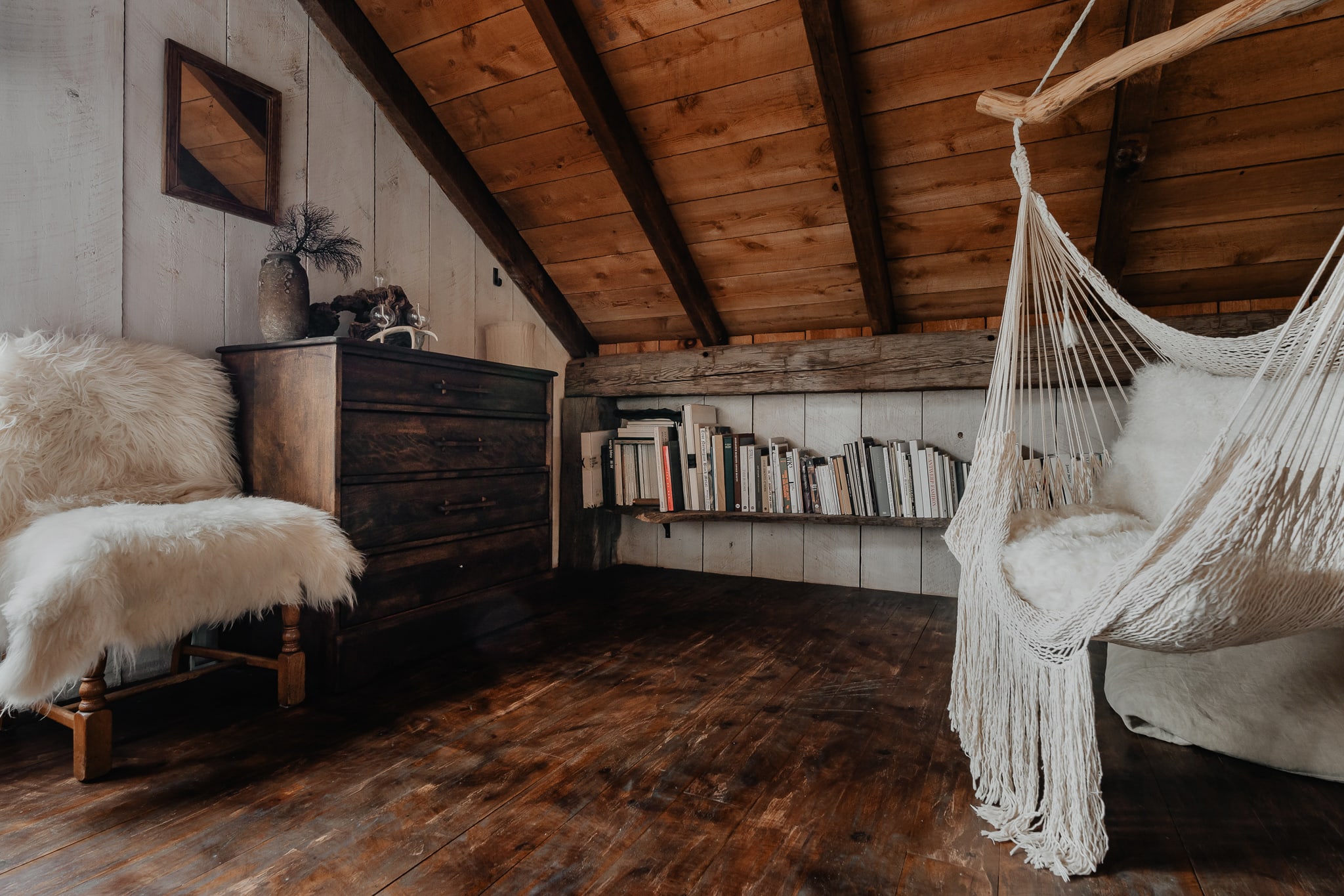 A rustic attic or loft space features sloped wooden plank ceilings and dark hardwood floors. A white woven hammock chair hangs near a bookshelf filled with books, and a wooden dresser with a furry chair completes the cozy nook.