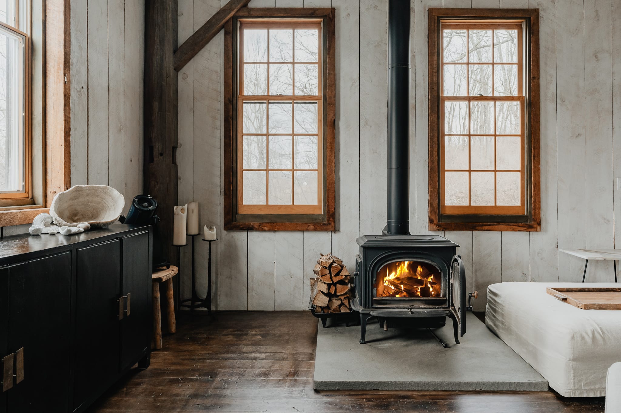 A cozy living area with white shiplap walls and dark hardwood floors features a black wood-burning stove on a concrete hearth, with stacked firewood beside it. Two large windows offer views of bare trees, and a light-colored sofa is partially visible.