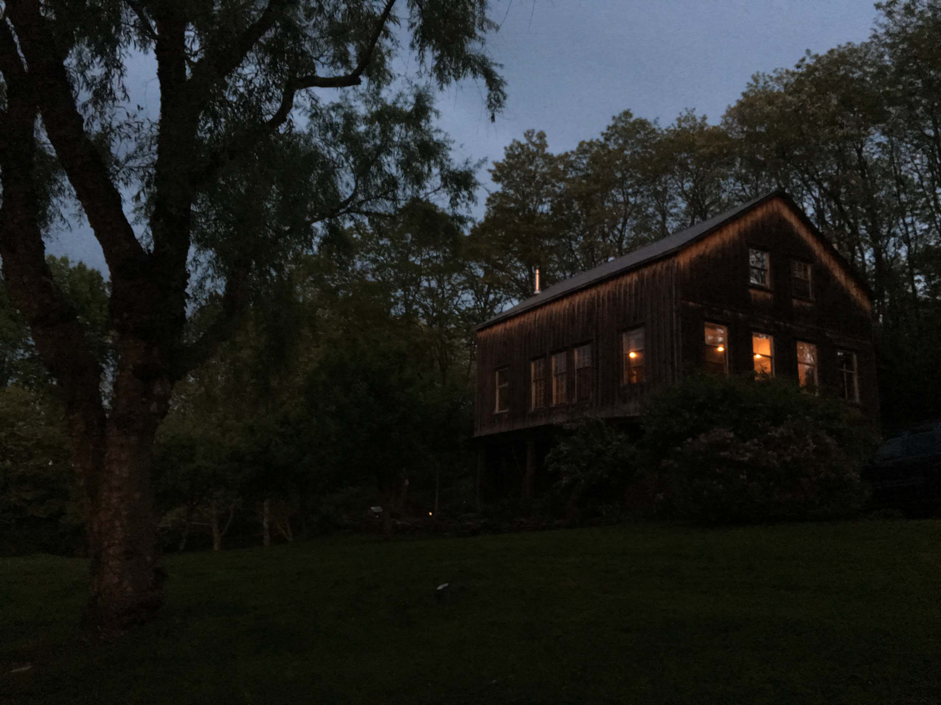 A rustic barn-style house with dark wooden siding and illuminated windows, captured at night under a dark sky. The surrounding trees and lawn are dimly lit, highlighting the warm glow emanating from the house.