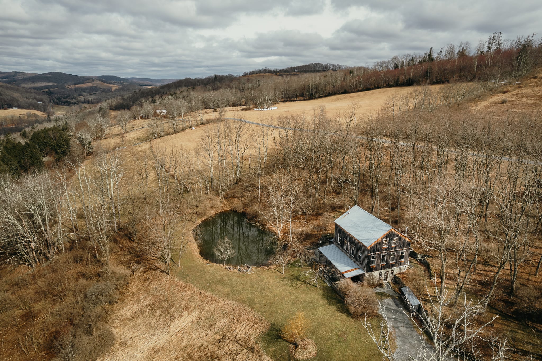 A wide aerial view of a rustic barn-style house and a dark pond, situated amidst a vast, rolling landscape of bare trees and dry fields under a cloudy sky. Distant hills complete the expansive rural scenery.