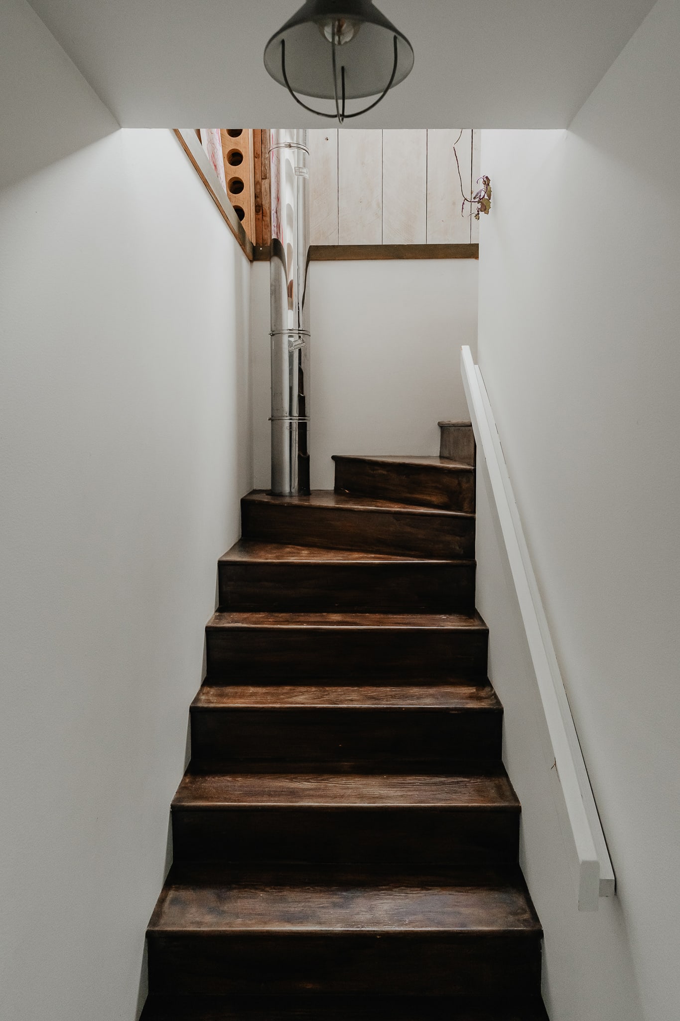 A narrow, dark wooden staircase with white walls and a simple white handrail, leading upwards. A metal pipe runs vertically alongside the stairs.