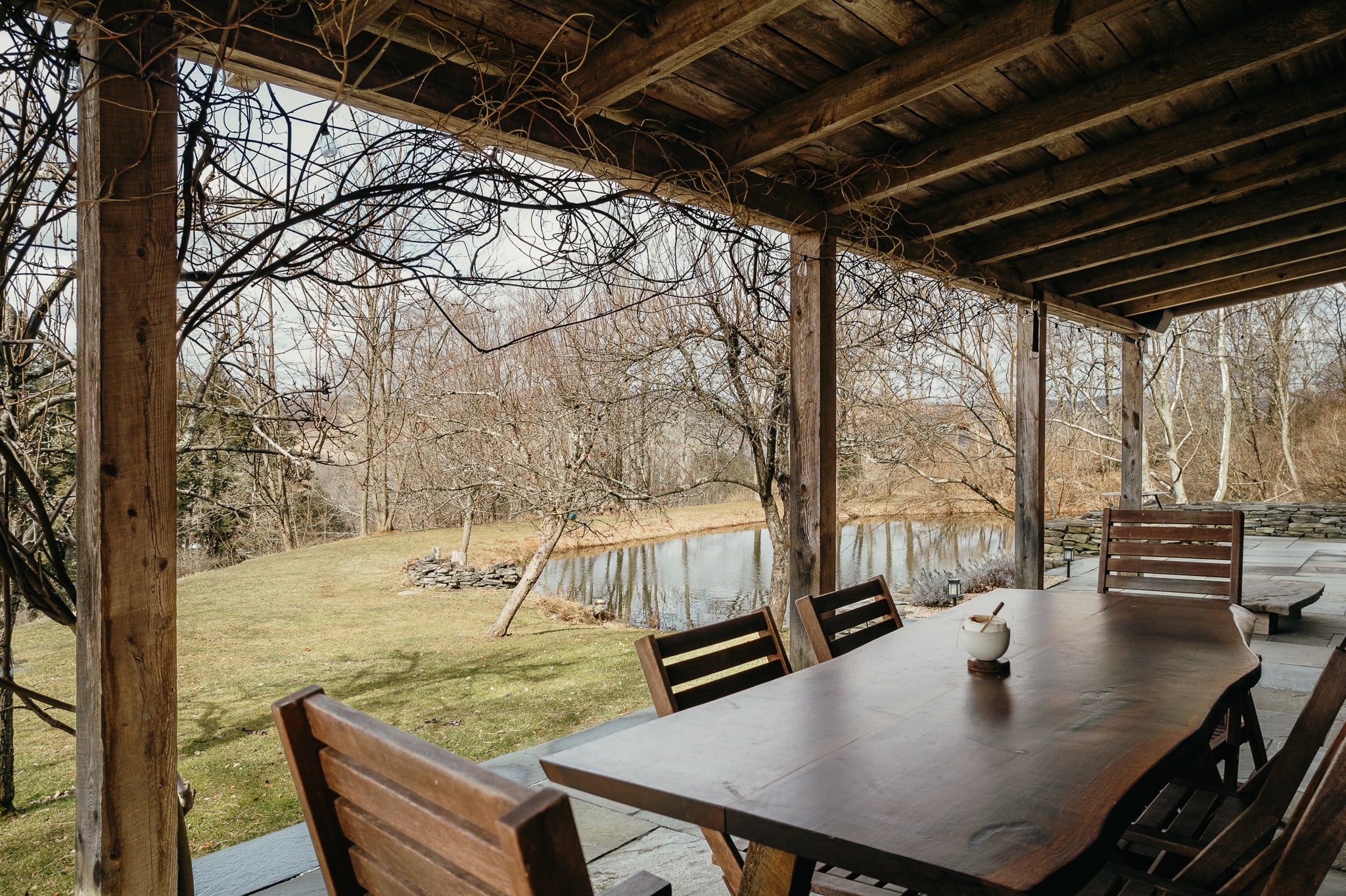 A covered outdoor porch features a large live-edge wooden dining table with matching chairs, set on a stone patio. Bare tree branches intertwine above, and a serene pond with a stone wall is visible through the trees in the background.