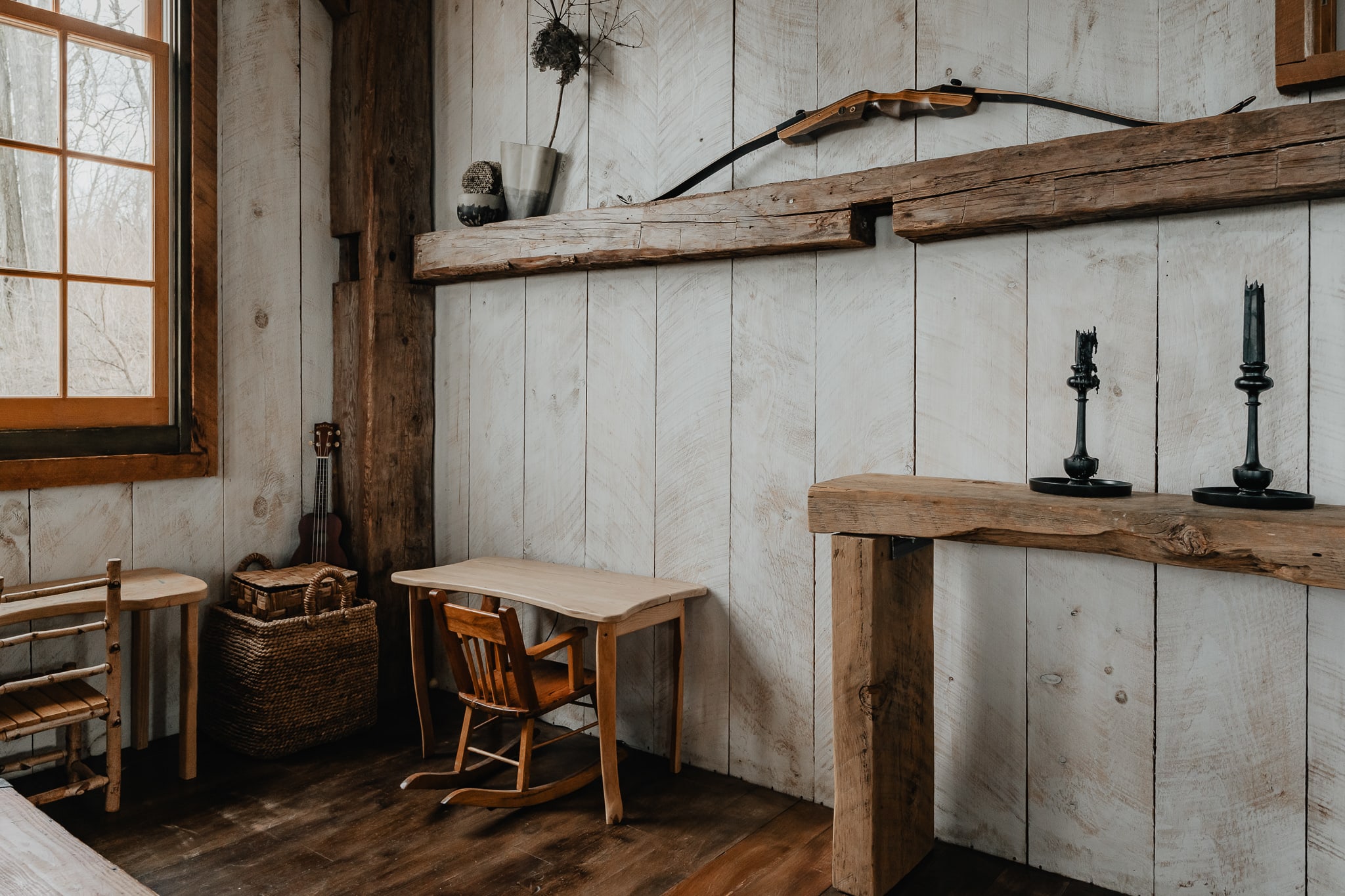 A rustic workspace area features a small wooden desk with a rocking chair, set against white shiplap walls and dark wood floors. A bow and arrows are displayed on a wooden beam above, and a guitar rests in a woven basket.