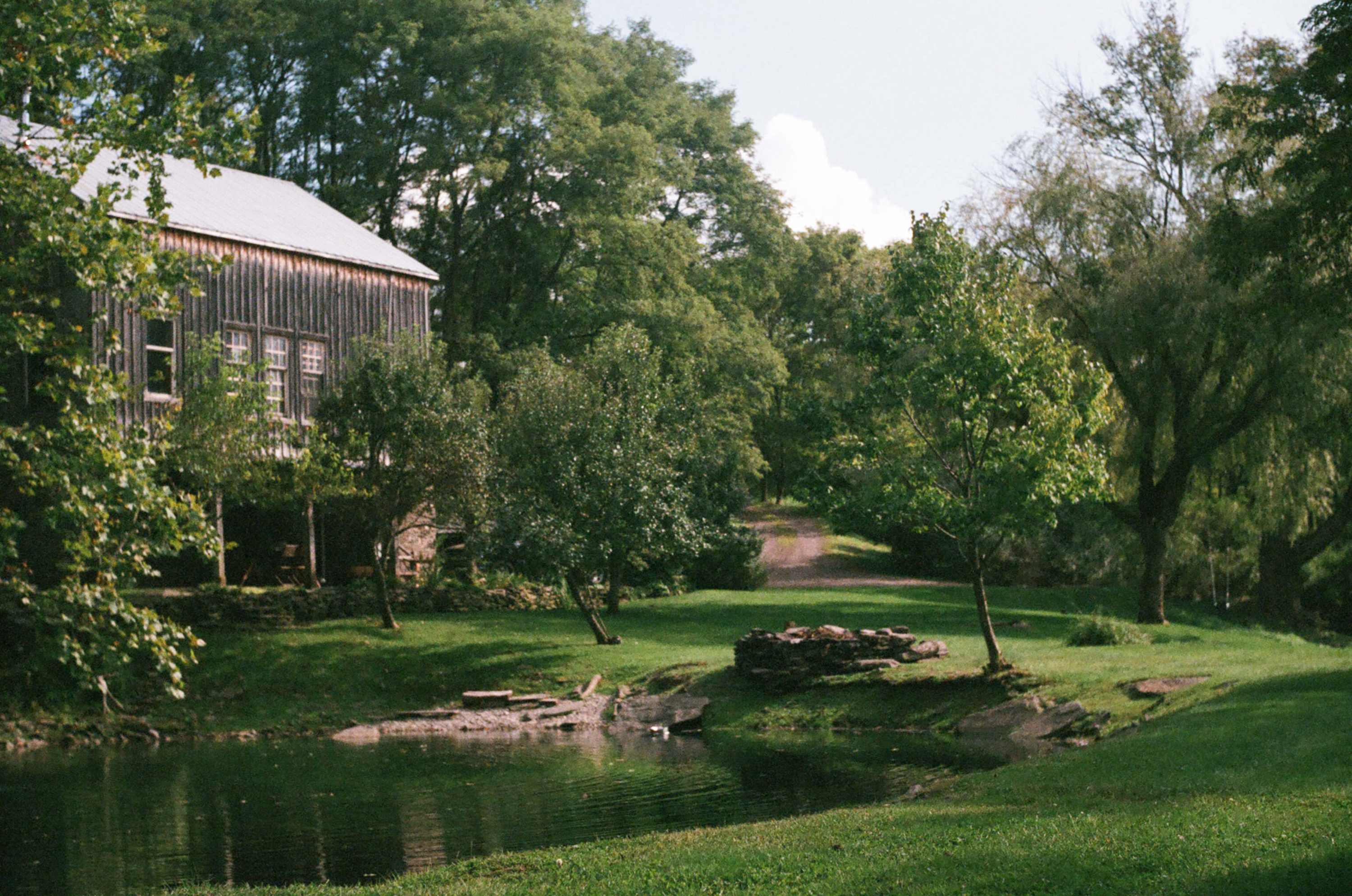 The rustic wooden barn-style house with a gray metal roof is seen from the back, surrounded by lush green grass and mature trees. A tranquil pond with a stone-lined edge is in the foreground, reflecting the greenery.