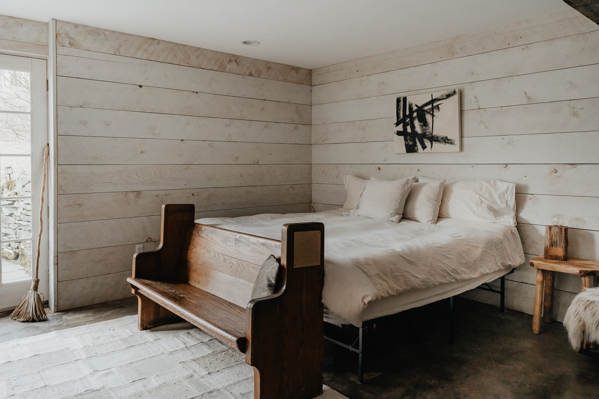 A rustic bedroom with white shiplap walls, a bed with a unique wooden church pew-style frame, and a large French door leading outside. A broom leans against the wall near the door.