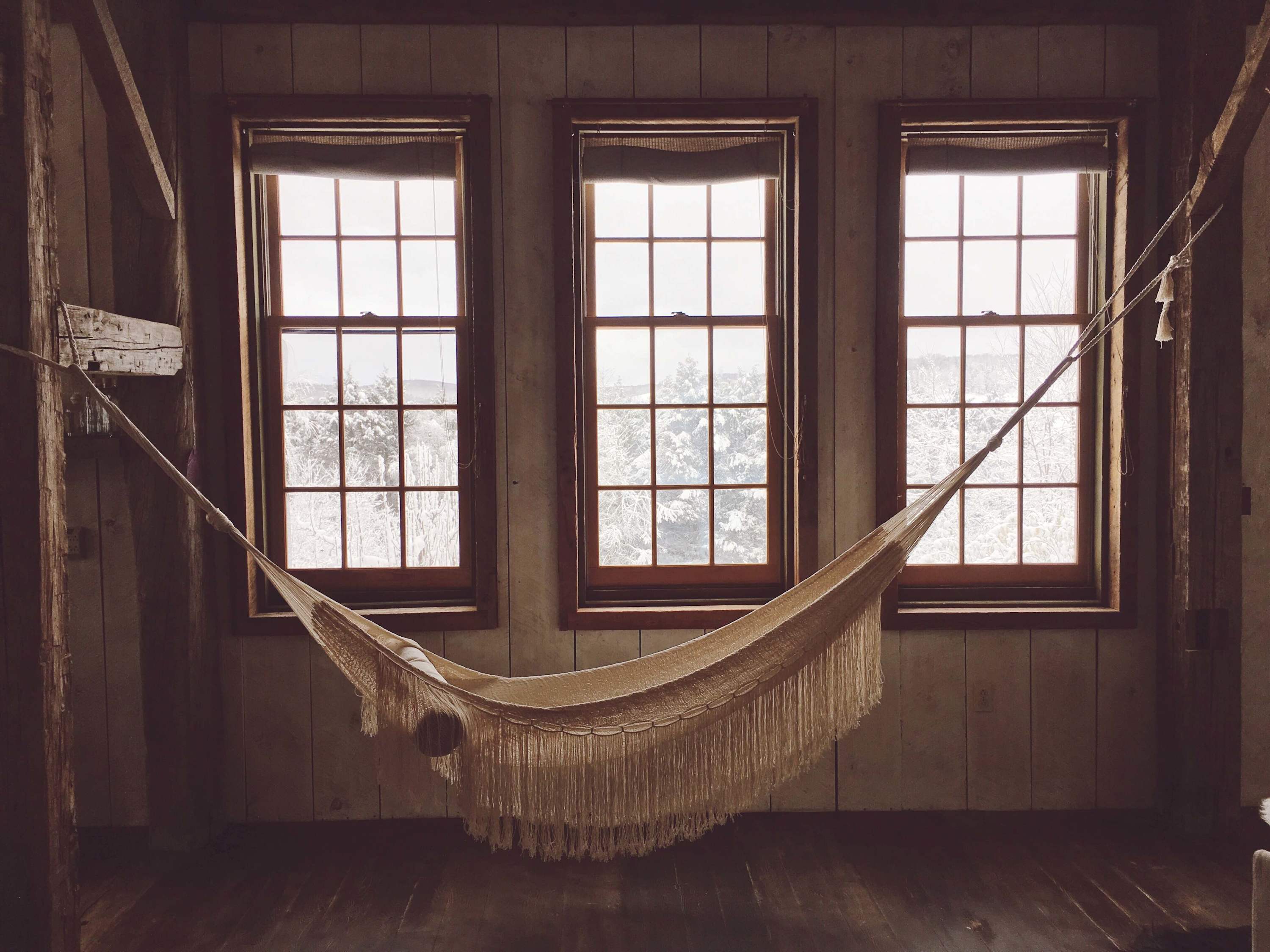 A cozy indoor hammock hangs in front of three large windows, offering a serene view of a snowy landscape. The room features rustic wood-paneled walls and dark wood floors.