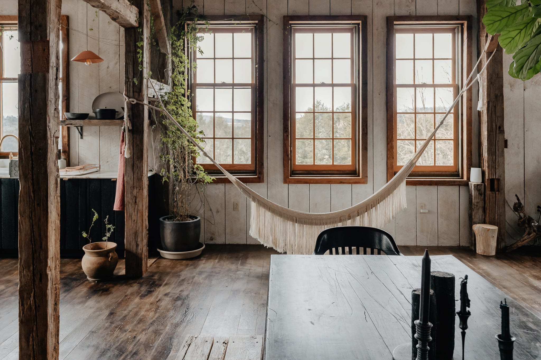 An inviting living space with dark hardwood floors and white shiplap walls features a woven hammock hanging by three large windows that overlook a wooded landscape. A large potted plant adds greenery, and a dark wooden table with candles is in the foreground.