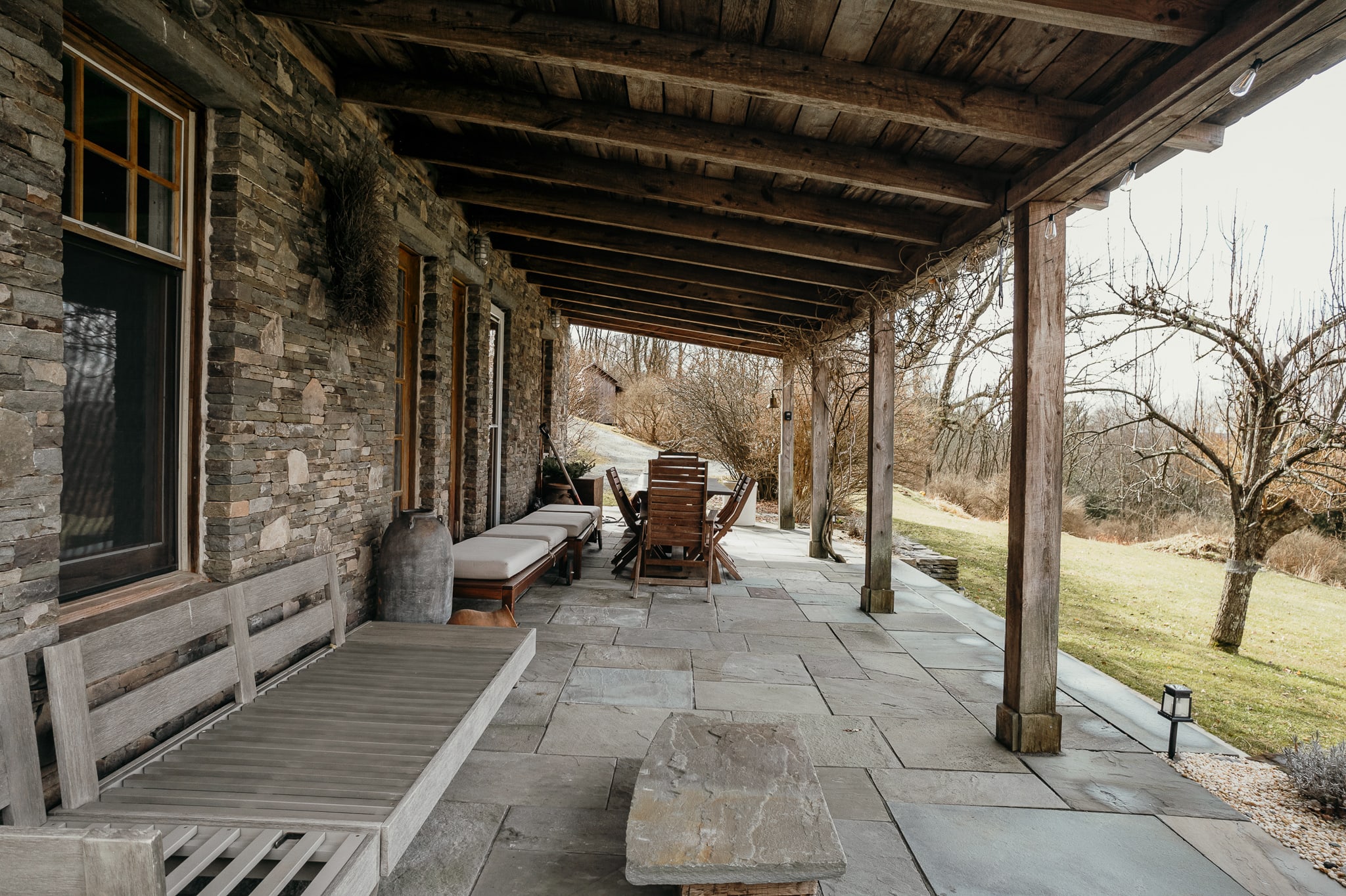A long, covered outdoor porch with a rustic wooden ceiling and a stone wall facade, featuring flagstone flooring and various wooden benches and chairs for outdoor seating. The porch overlooks a grassy area with trees.