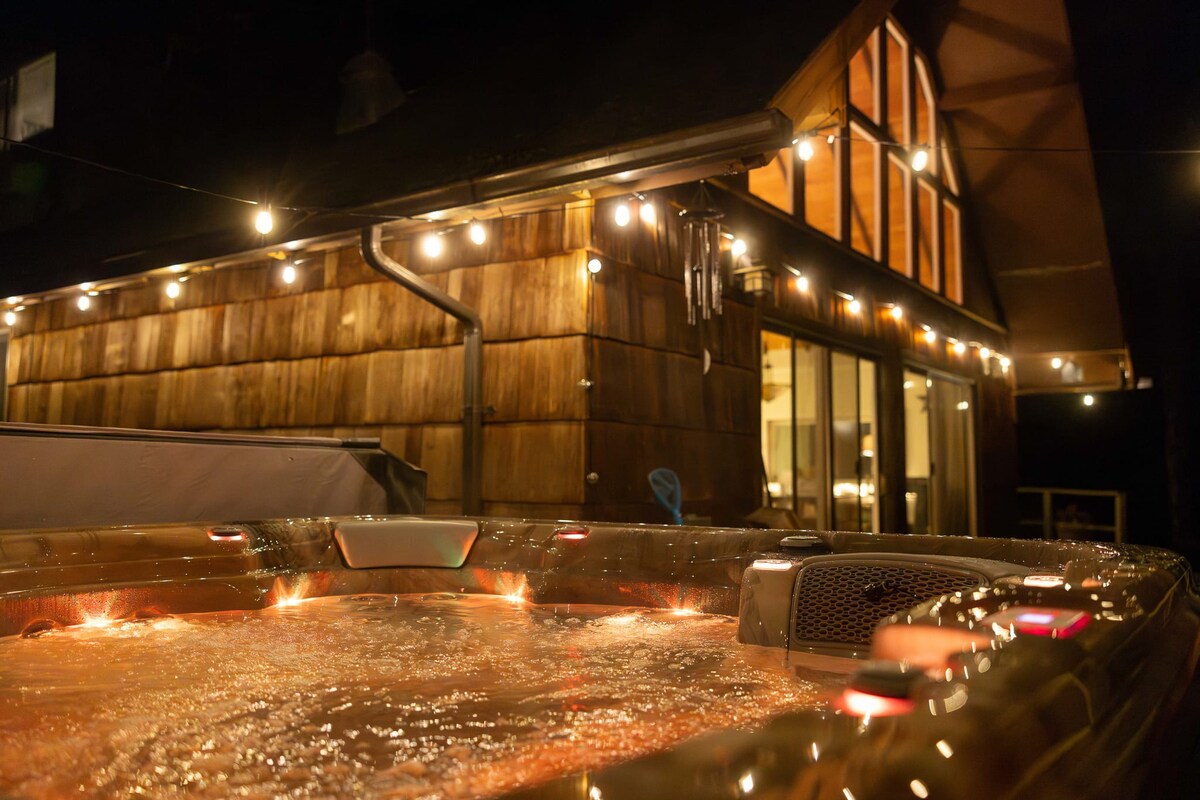 Nighttime view of the illuminated hot tub on the deck, with string lights adding a festive ambiance to the rustic cabin exterior. The interior lights of the cabin are visible through the windows.