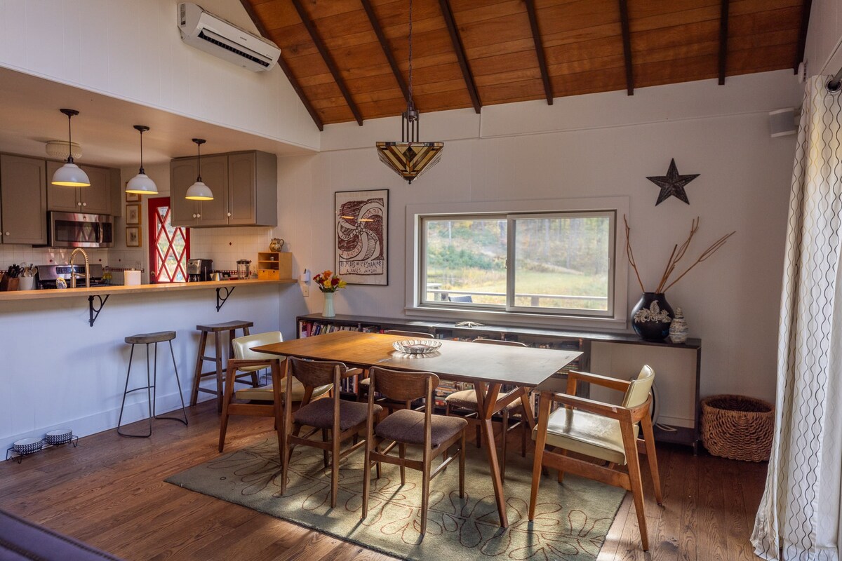 Open-concept dining area featuring a wooden dining table and chairs, with a kitchen breakfast bar and white pendant lights visible in the background, under a vaulted wood-paneled ceiling.