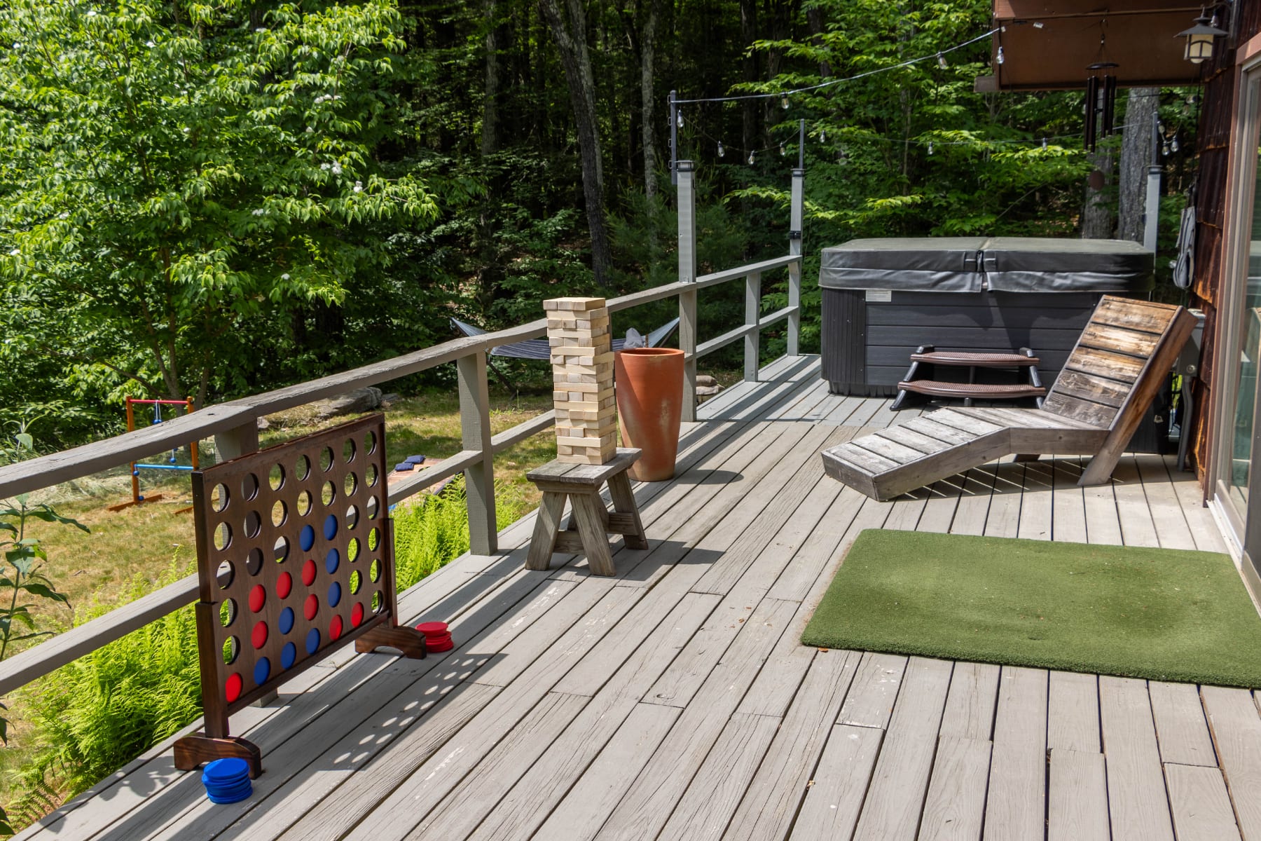Expansive wooden deck featuring a hot tub, a wooden lounge chair, and a giant Connect Four game. The deck is surrounded by a railing and overlooks a grassy area with trees.