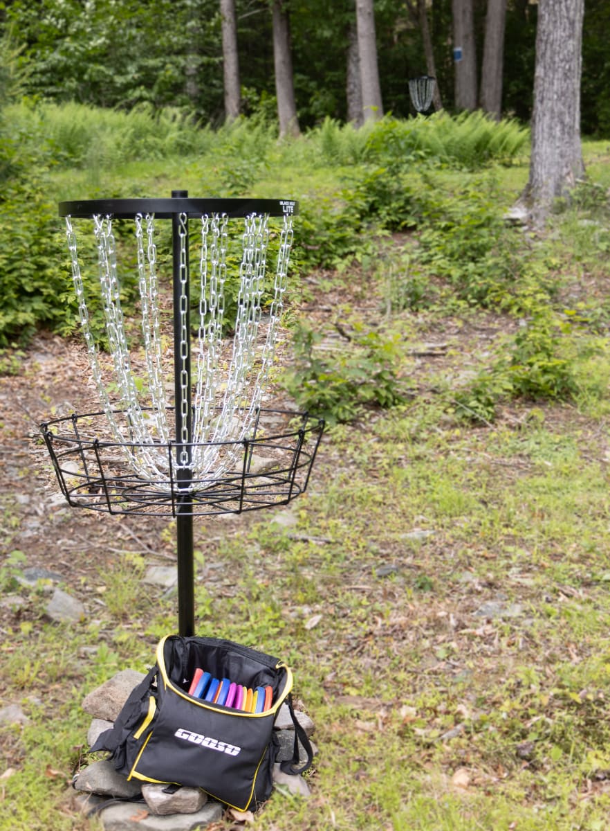 A black metal disc golf basket with silver chains stands in a grassy, wooded area, with a black bag filled with colorful discs at its base. Another disc golf basket is visible in the distance among the trees.