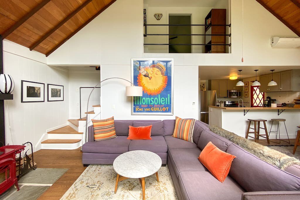 Cozy living room with a large purple sectional sofa and a round marble coffee table on an area rug, featuring a vaulted wood-paneled ceiling and a glimpse of a loft area above.
