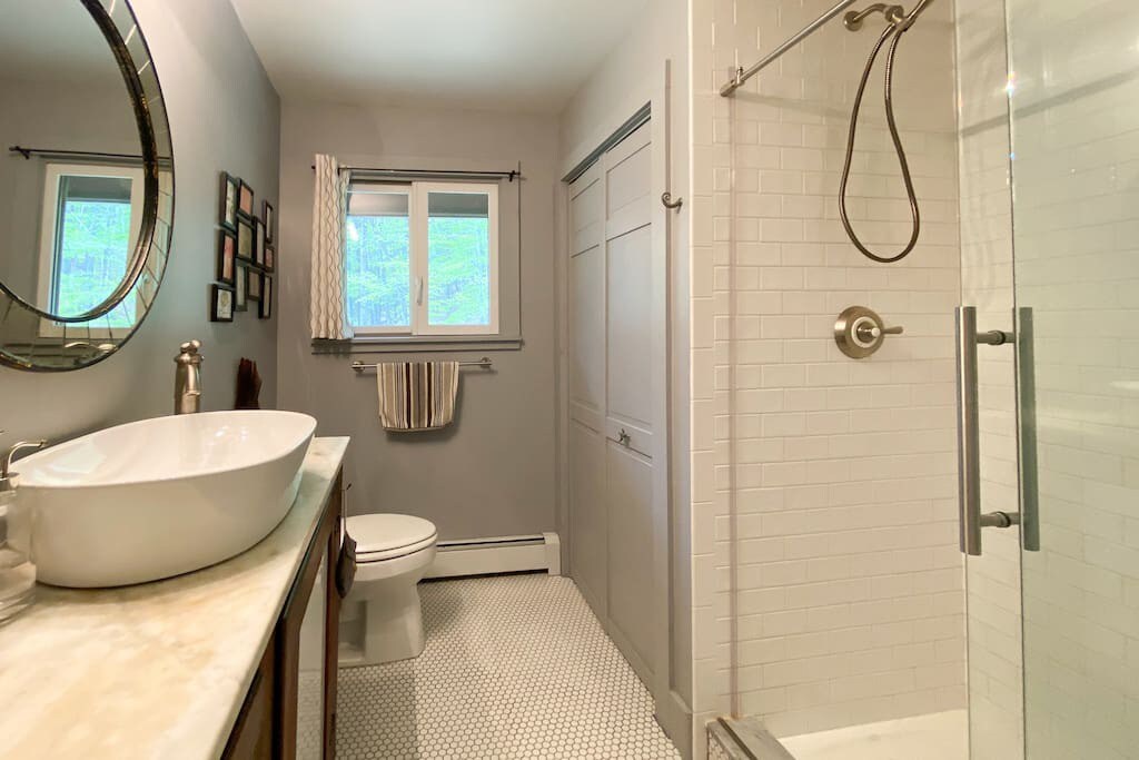 Modern bathroom with a white vessel sink on a marble-topped vanity, a toilet, and a walk-in shower with white subway tiles and a glass door. The floor is covered in small white hexagonal tiles.