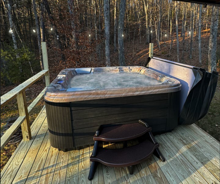 Outdoor hot tub with bubbling water, situated on a wooden deck surrounded by a railing and string lights. The background shows a dense forest under a clear sky.