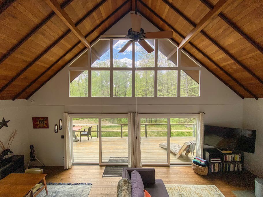 Bright living room with a high vaulted wooden ceiling and a wall of large windows offering views of the surrounding green forest. Sliding glass doors lead to an outdoor deck.
