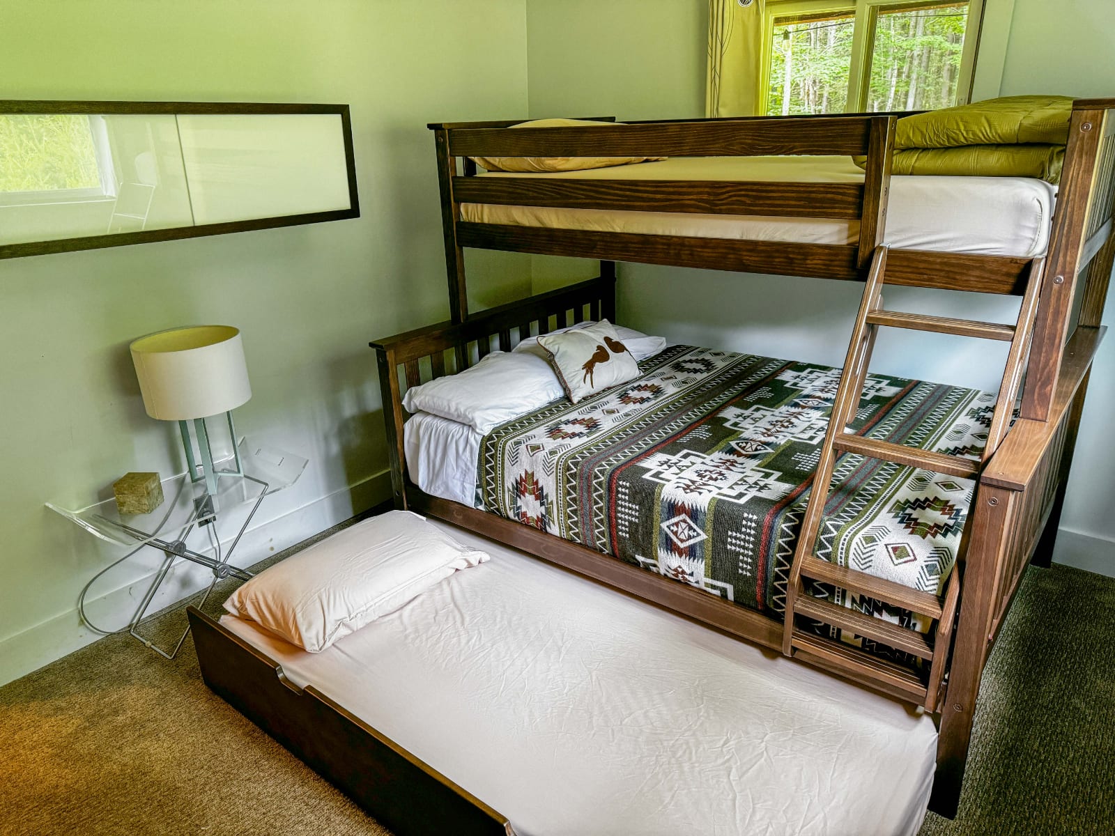 Cozy bedroom featuring a dark wood bunk bed with a trundle bed underneath, adorned with a patterned blanket and a deer-themed pillow. A modern lamp sits on a clear acrylic side table next to the bed.