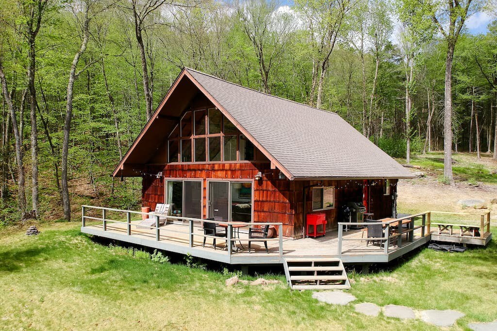 Chalet-style house with warm wooden siding and an A-frame roof, featuring a large wraparound deck with outdoor seating and a grill, nestled among lush green trees under a bright sky.
