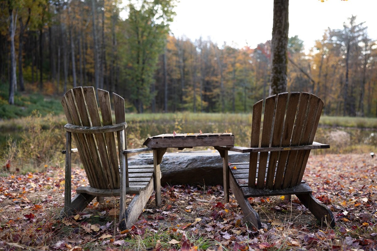 Two rustic wooden Adirondack chairs with a small table between them, facing a serene pond. The ground is covered with colorful autumn leaves, and a forest is visible in the distance.