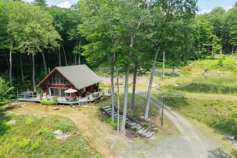 Aerial view of the A-frame cabin with a large deck, situated on a grassy hill next to a pond, surrounded by dense green forest. A gravel driveway leads up to the property.